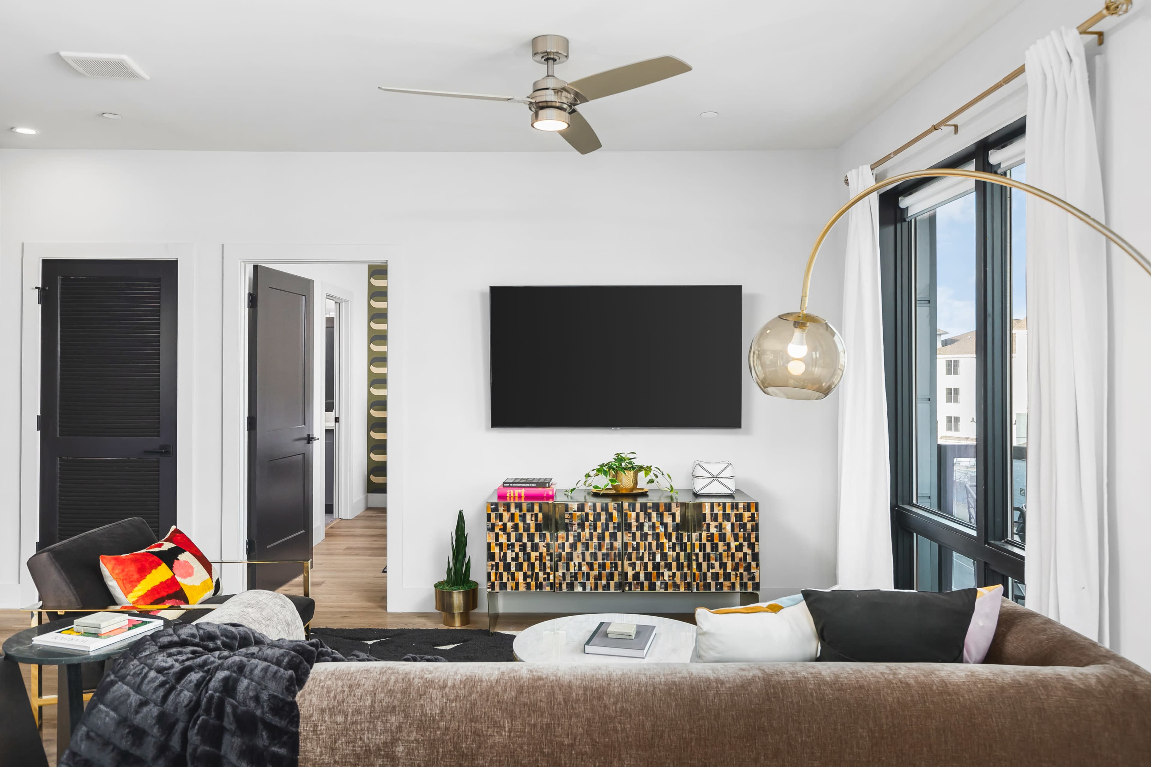 A modern living room features a sectional couch, a contemporary coffee table, and a wall-mounted television above a patterned console table, with large windows and a doorway leading to another room.