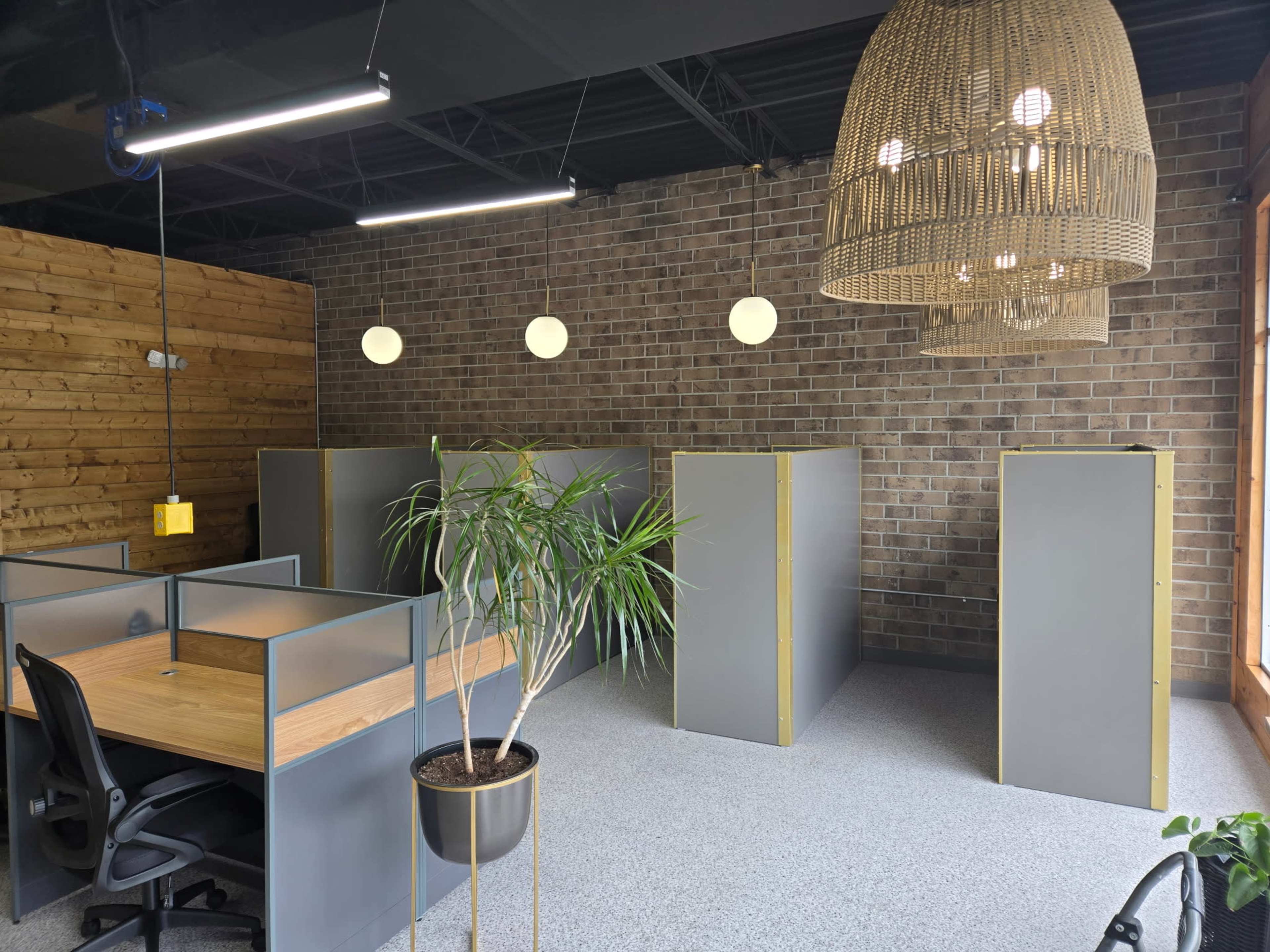 The image shows a modern office space with gray workstations separated by low partitions, a potted plant, and pendant lighting against a brick wall.