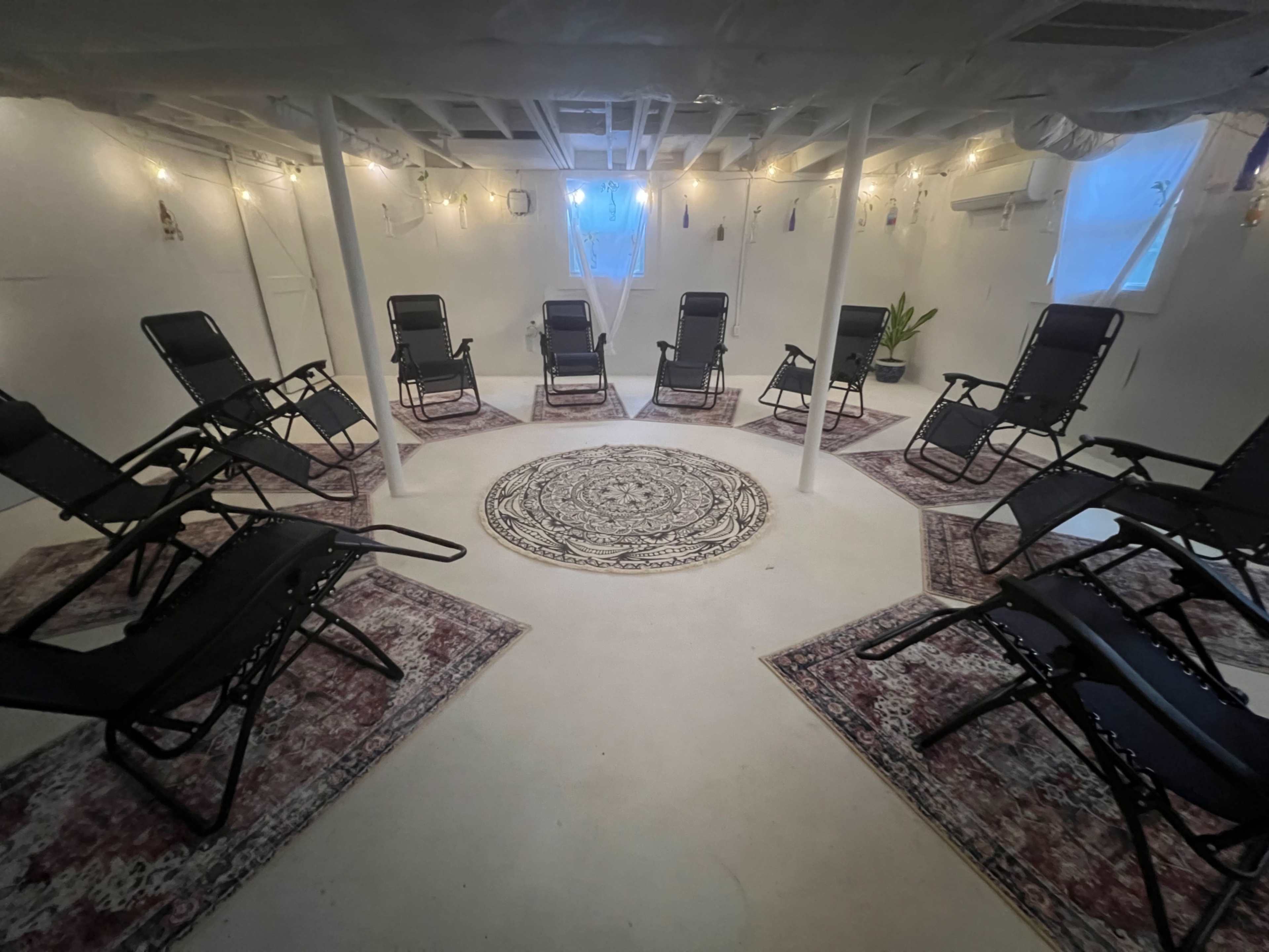 A circular seating arrangement with black reclining chairs is set on a light-colored floor adorned with a central rug in a well-lit basement space.