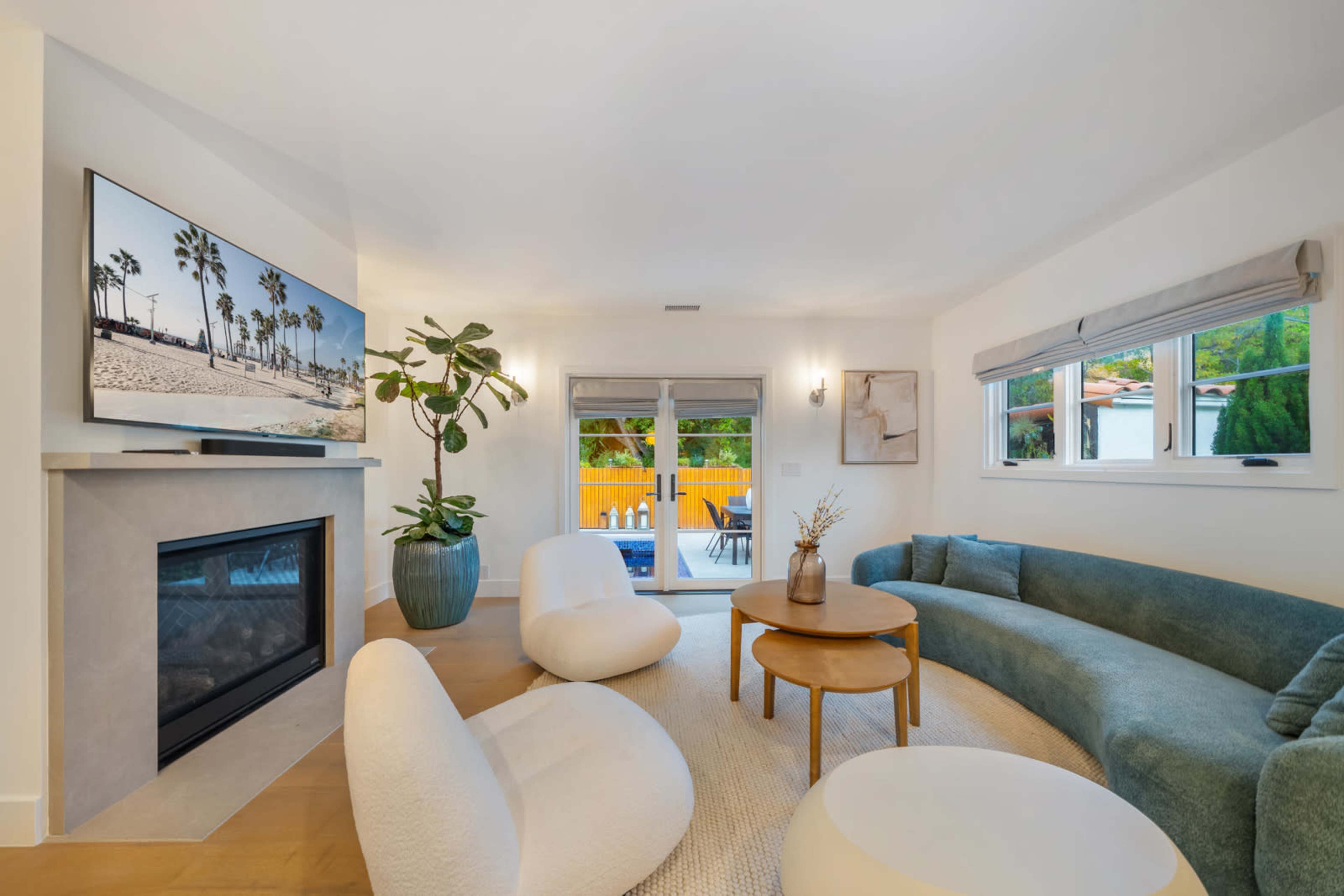 The living room features a curved sofa, two rounded chairs, a round coffee table, a fireplace, and a large TV mounted on the wall, with sliding glass doors leading to an outdoor area.