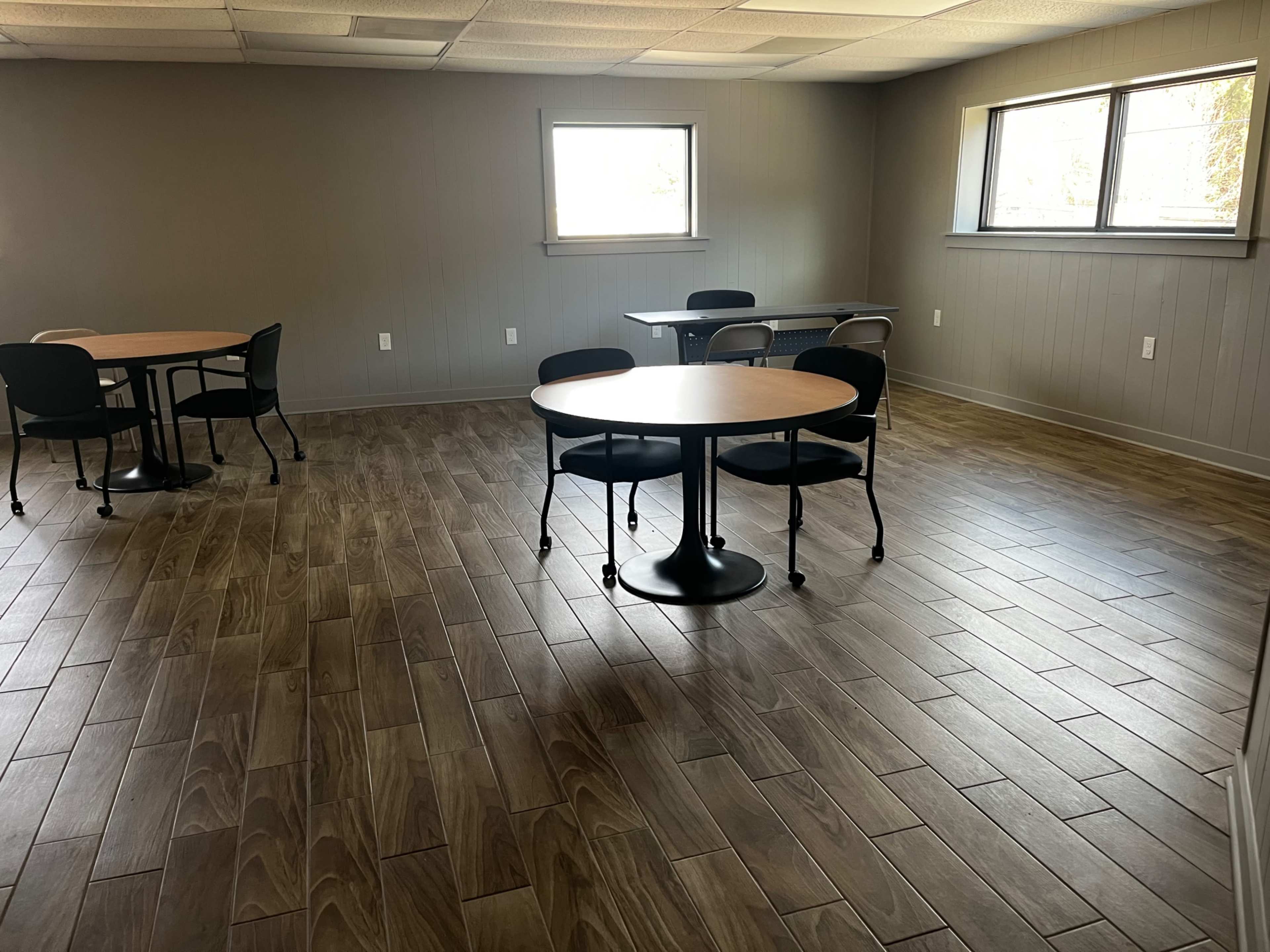 The image shows a spacious room with two circular tables and black chairs, illuminated by natural light from windows and featuring wooden tiled flooring.