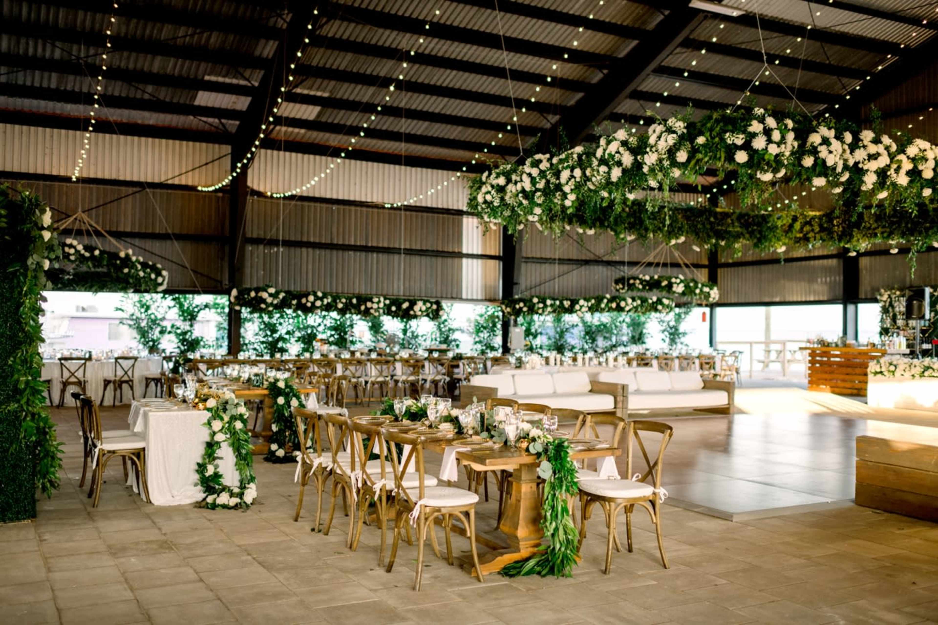 A beautifully decorated event space with wooden tables, greenery, and floral arrangements, set under a large metal structure.
