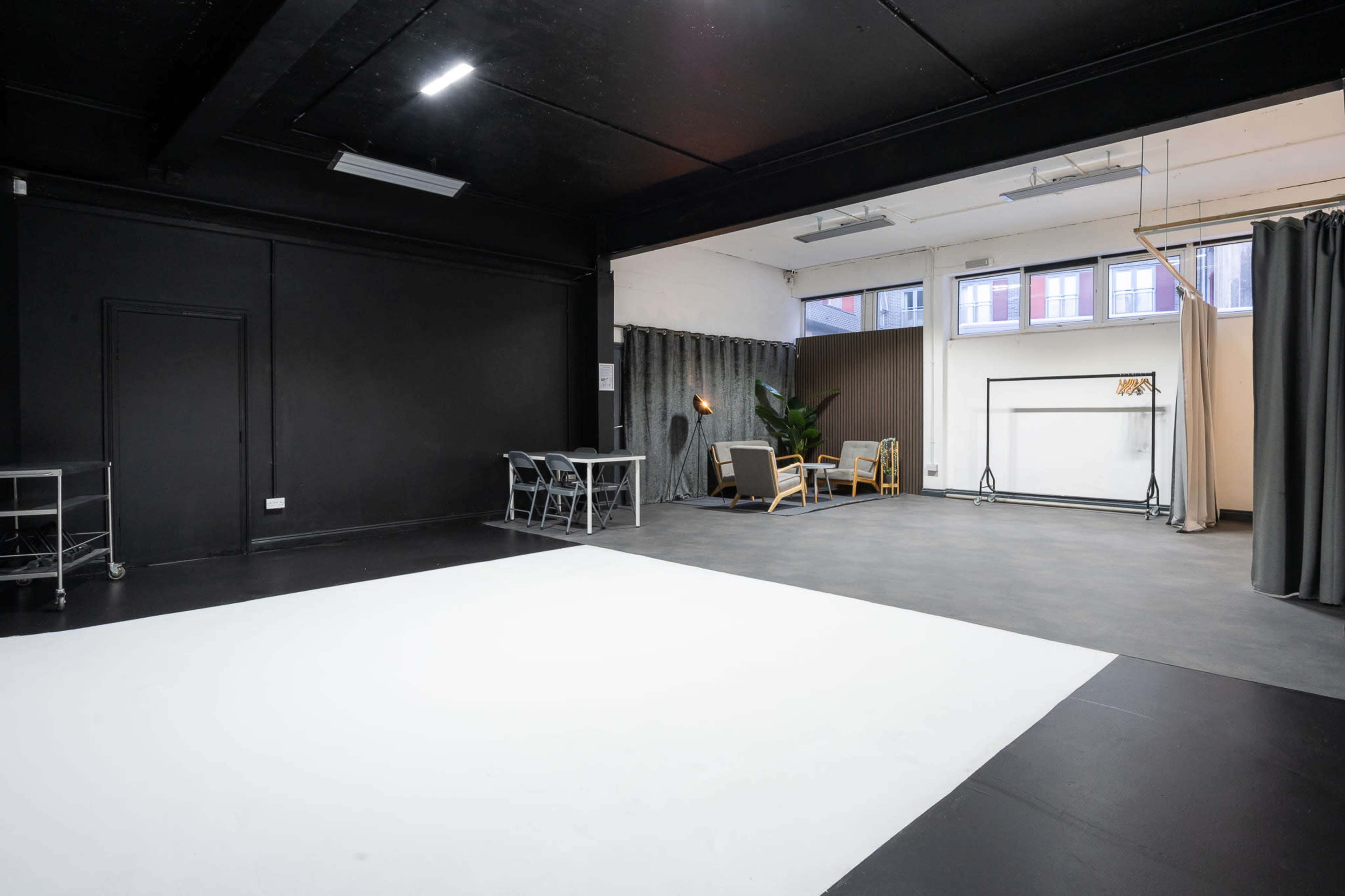 The image shows a spacious studio with a black ceiling and walls, featuring a white area on the floor, a table with chairs, and a seating area with a plant in the background.