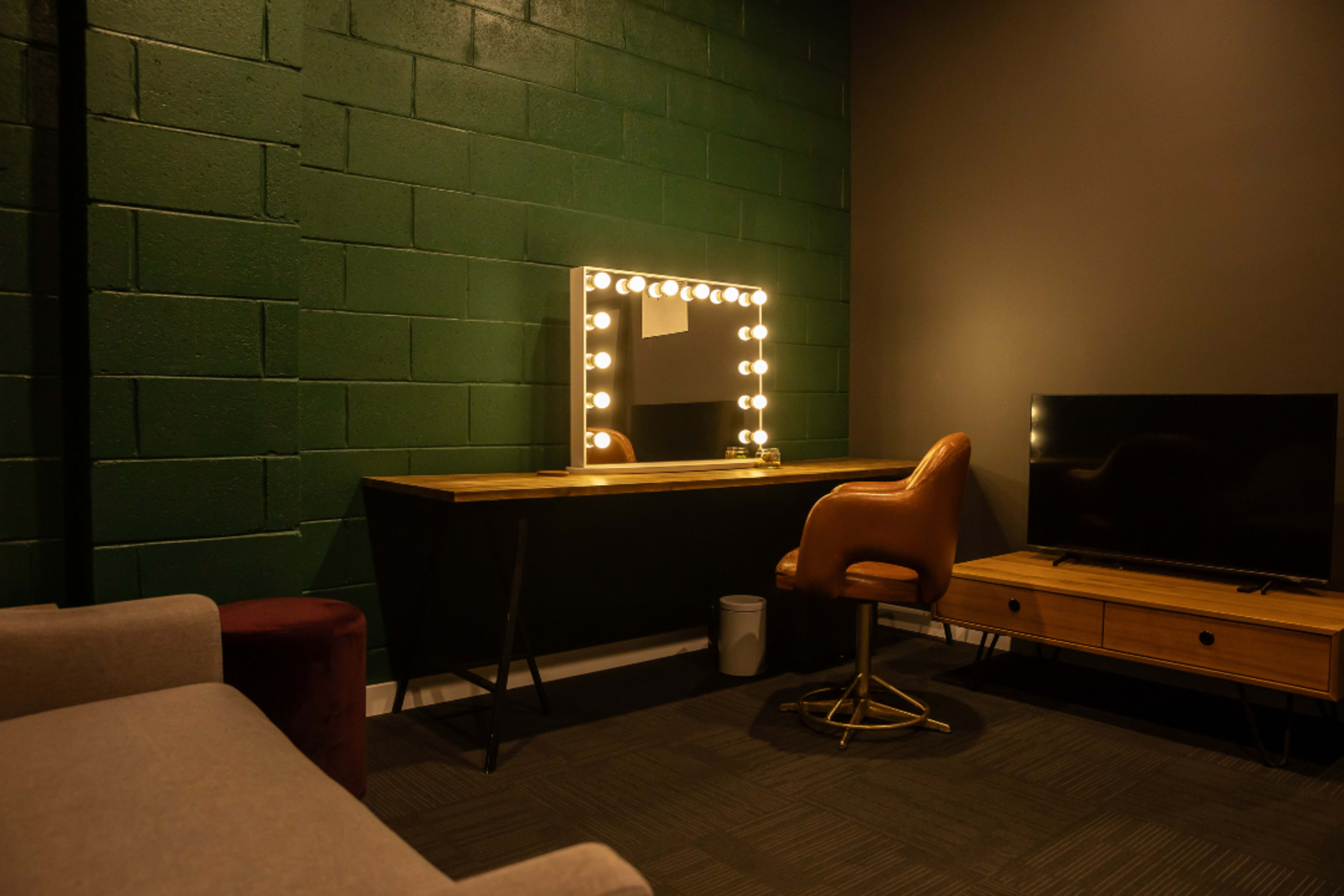 The image shows a room with a green brick wall featuring a large illuminated mirror above a wooden vanity, a brown chair, and a television on a low wooden cabinet.