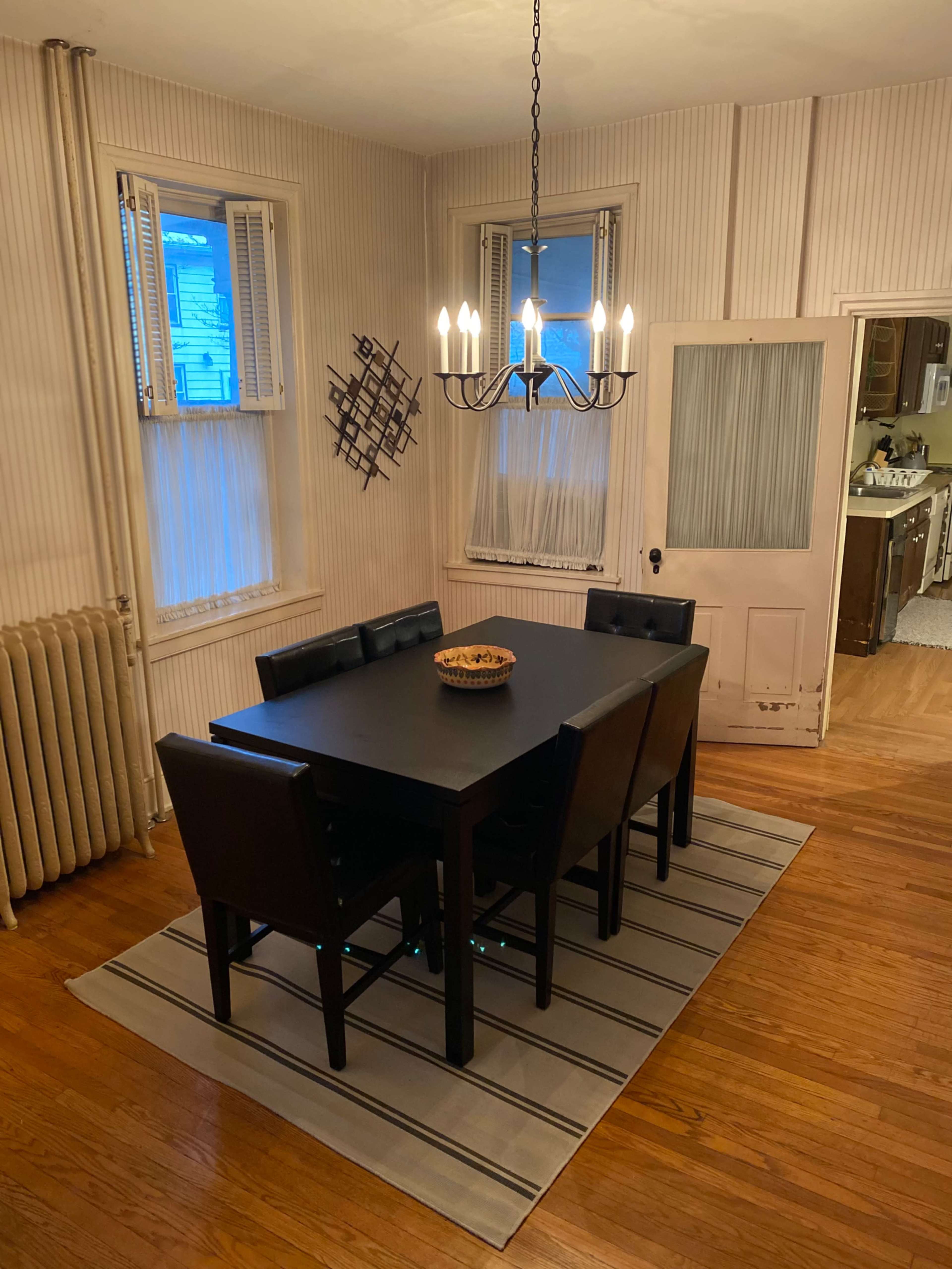 A black dining table surrounded by six chairs is situated on a striped rug in a room with wood flooring and light-colored walls, featuring a chandelier overhead and two windows.
