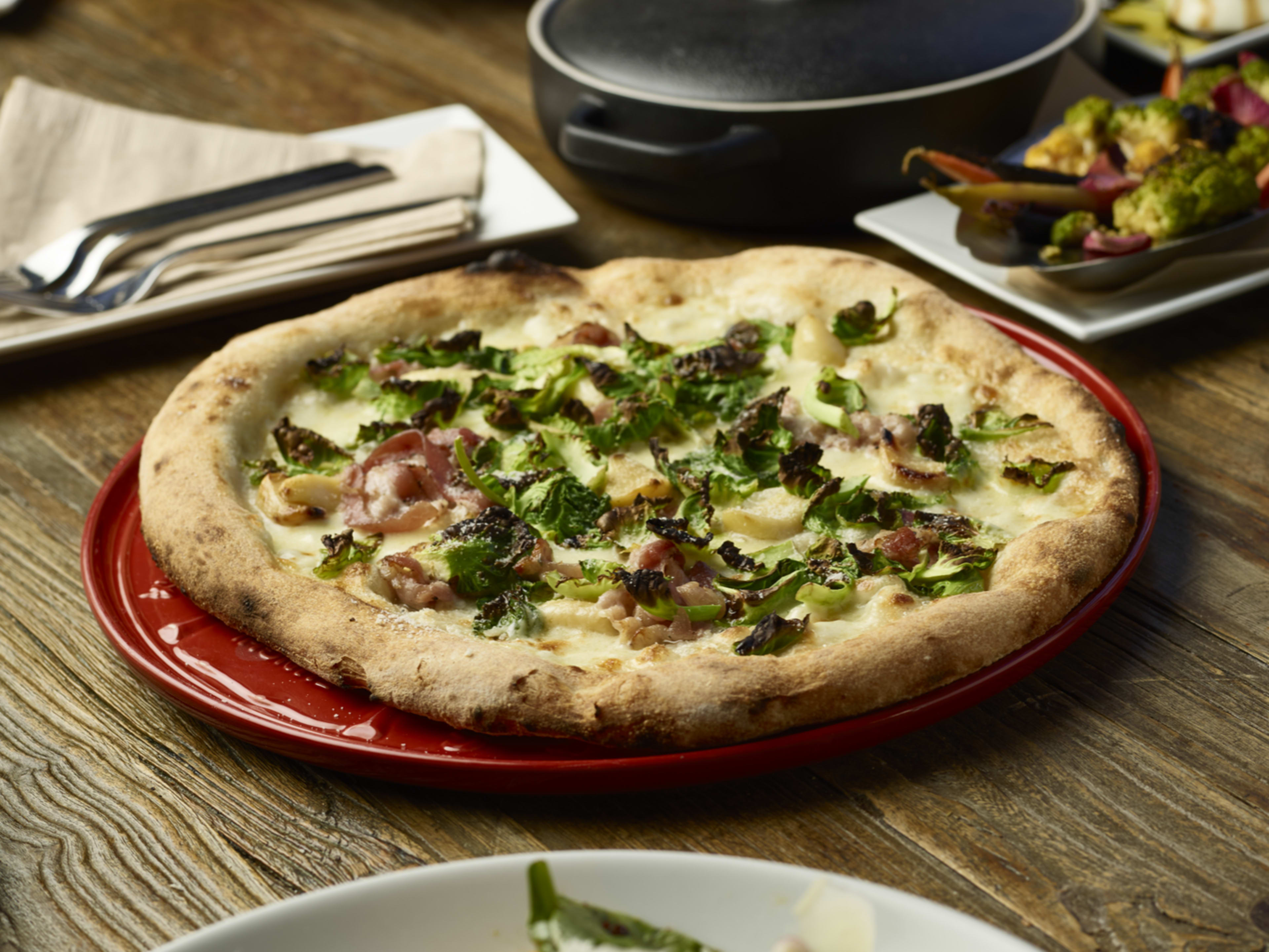 A pizza topped with greens and slices of meat is placed on a red dish on a wooden table alongside utensils and other dishes.