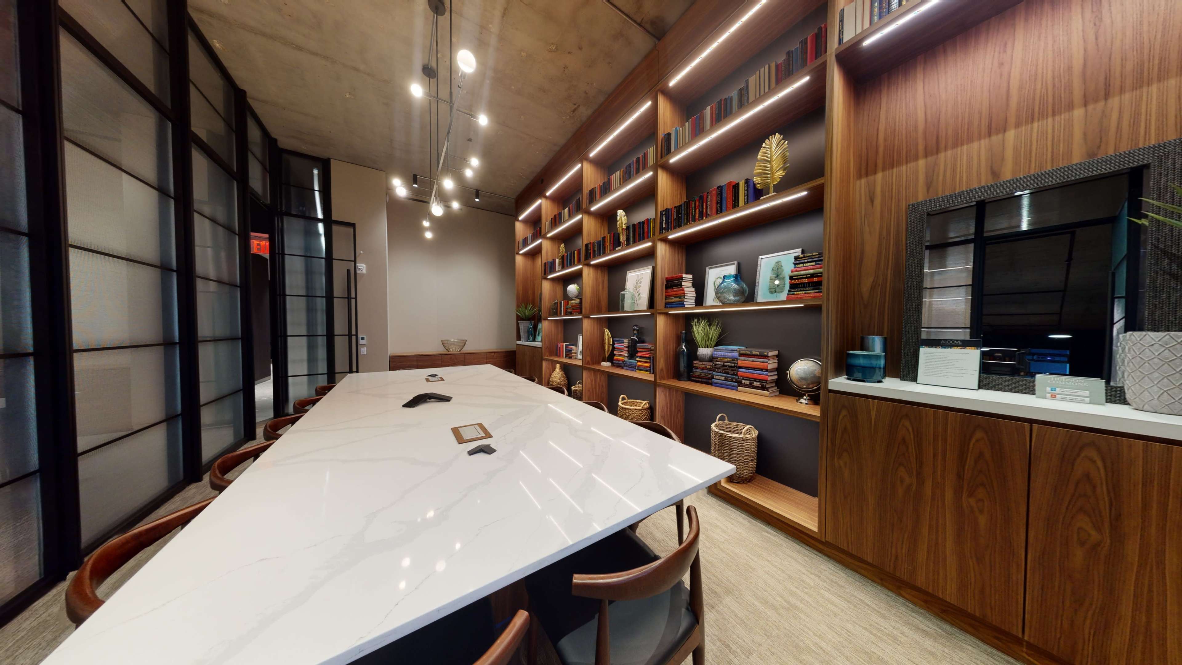 The image shows a modern conference room with a large marble table, wooden shelving filled with books and decorative items, and a wall of glass panels.