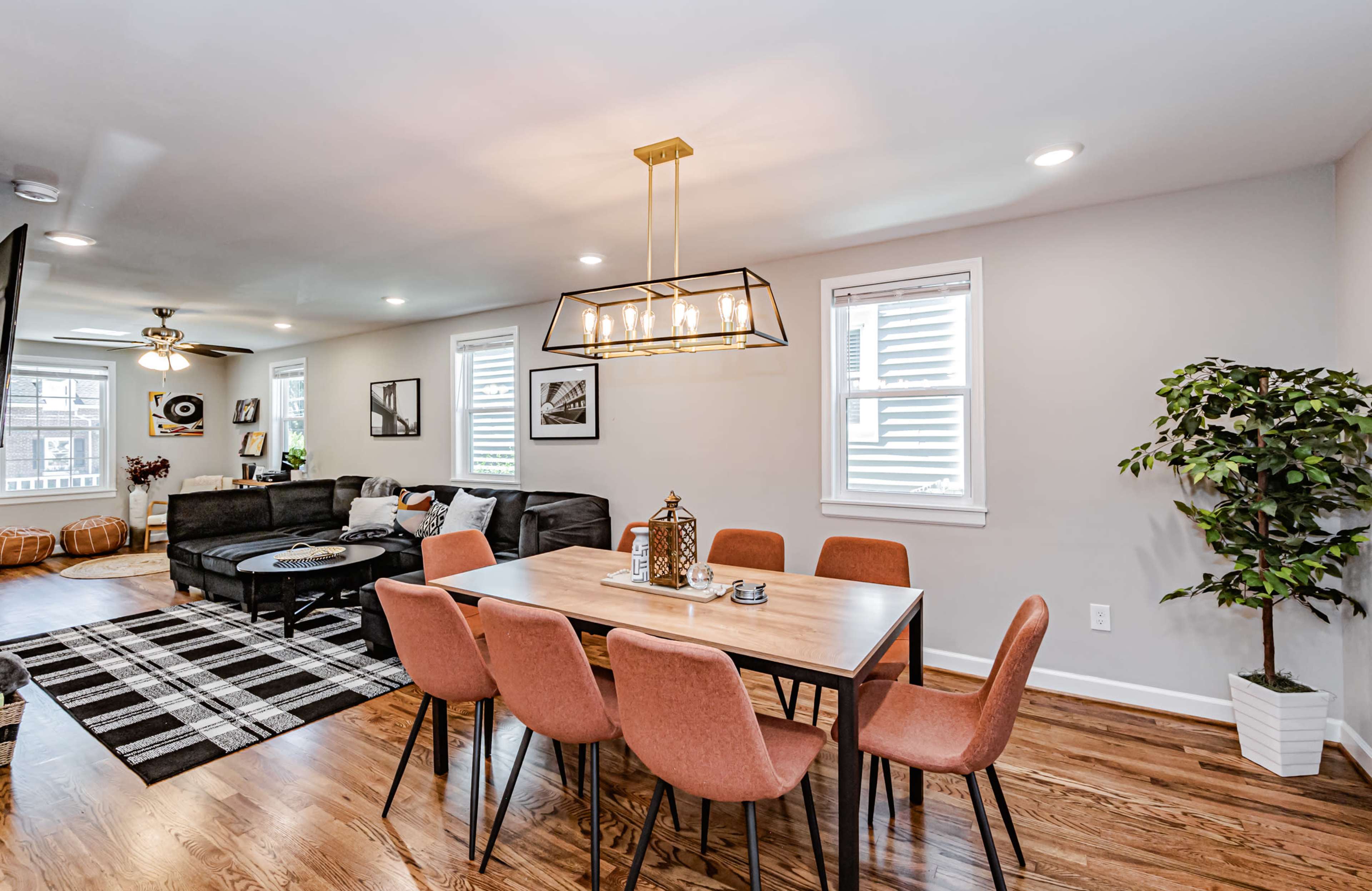 A modern dining area features a wooden table surrounded by eight pink chairs, with a black sectional couch and decorative plants in a well-lit, open space.