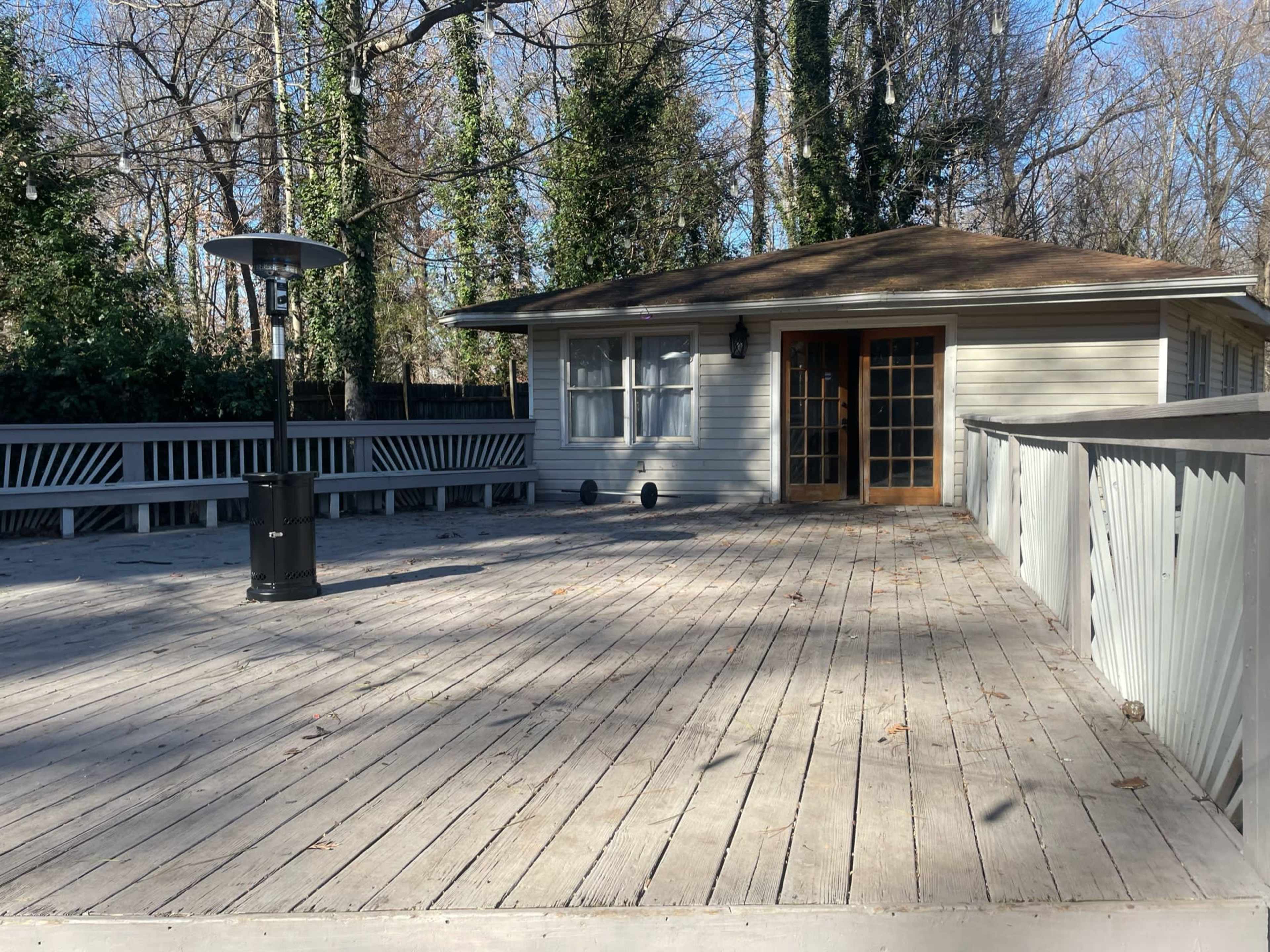 The image shows a wooden deck with a patio heater, surrounded by trees, adjacent to a house with double doors.