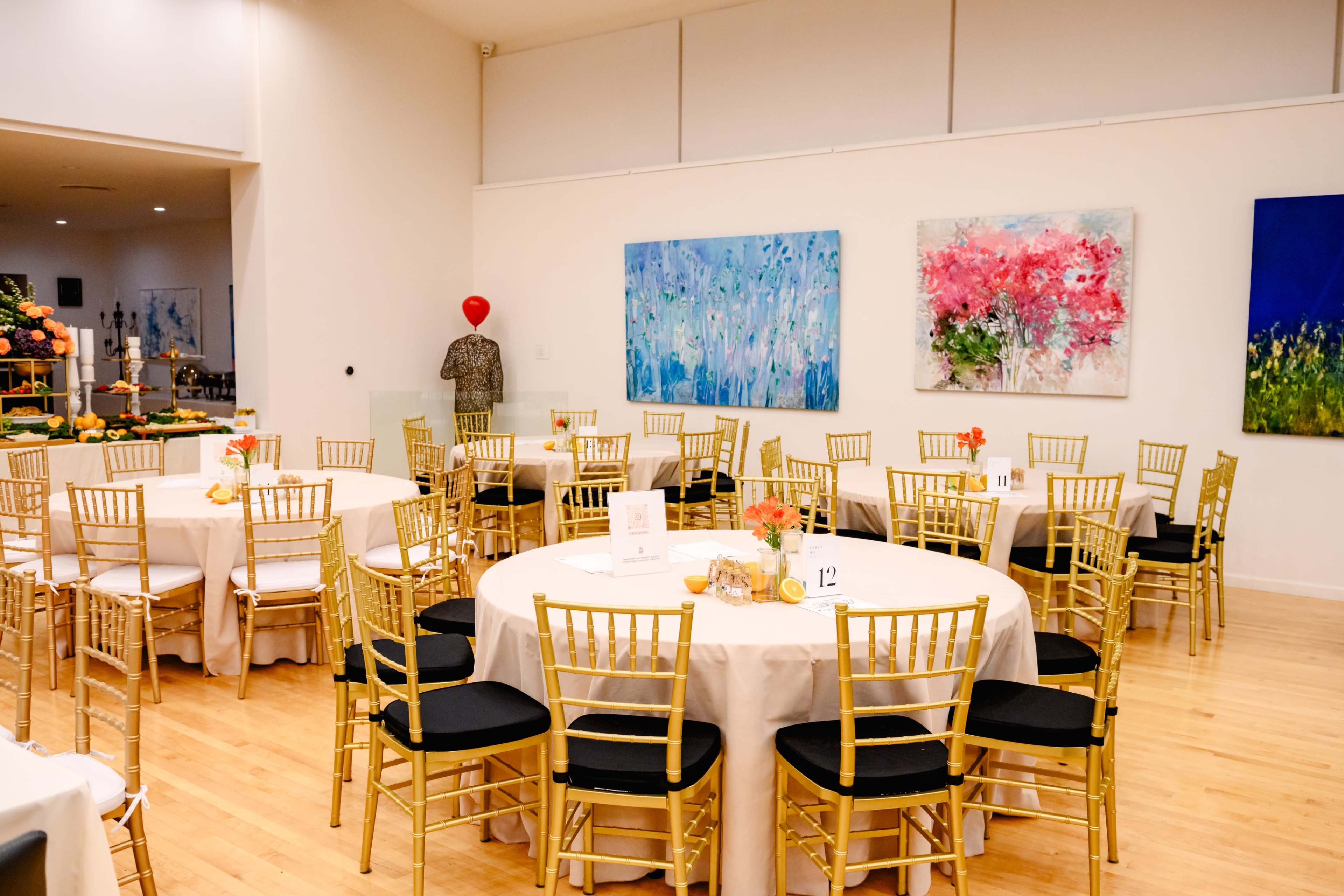 The image shows a brightly lit event space with round tables covered in white tablecloths, gold chiavari chairs, and colorful artwork on the walls.