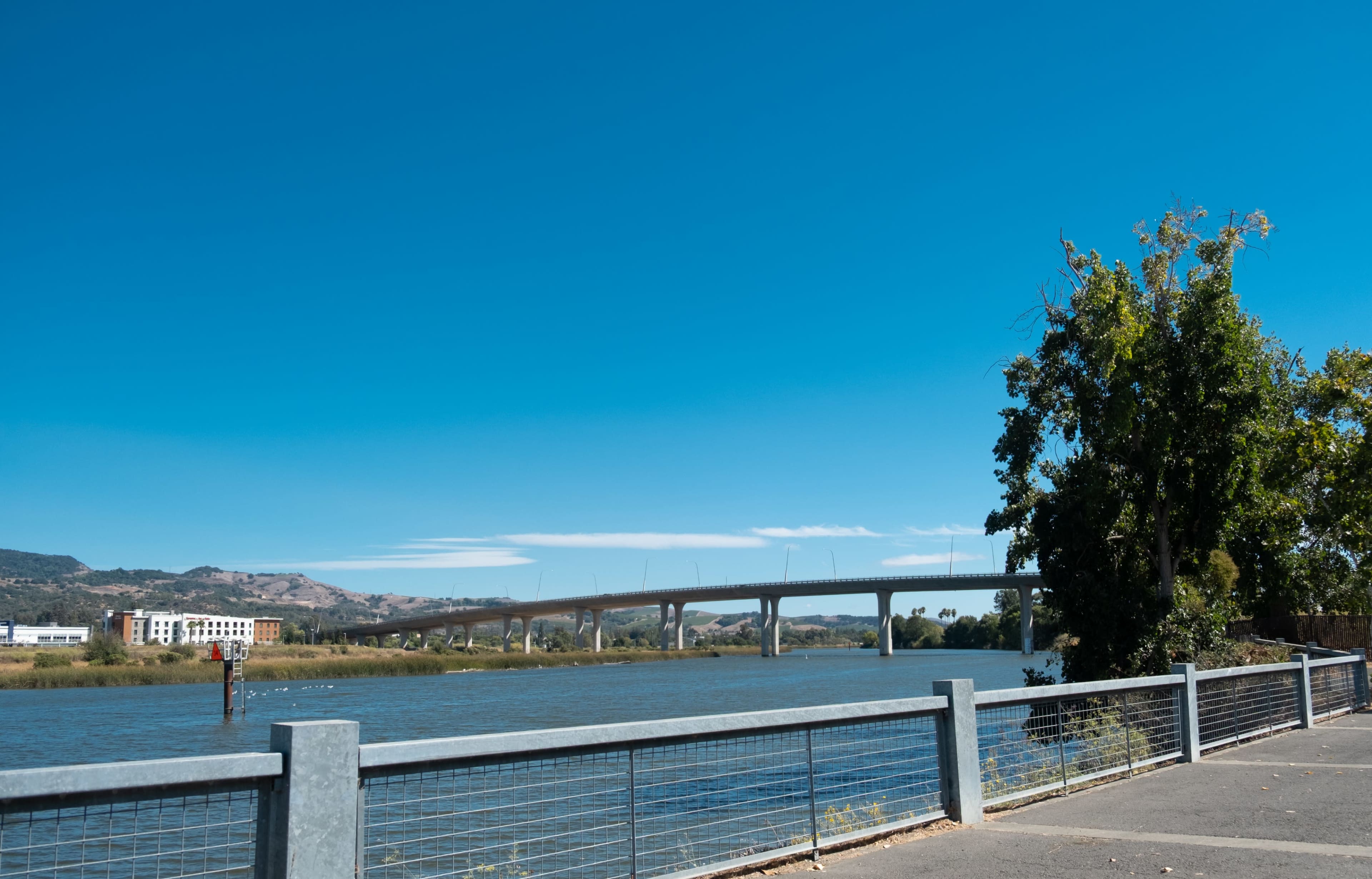 A concrete walkway lined with a metal railing runs alongside a river, with a bridge arching over the water and hills visible in the background under a clear blue sky.