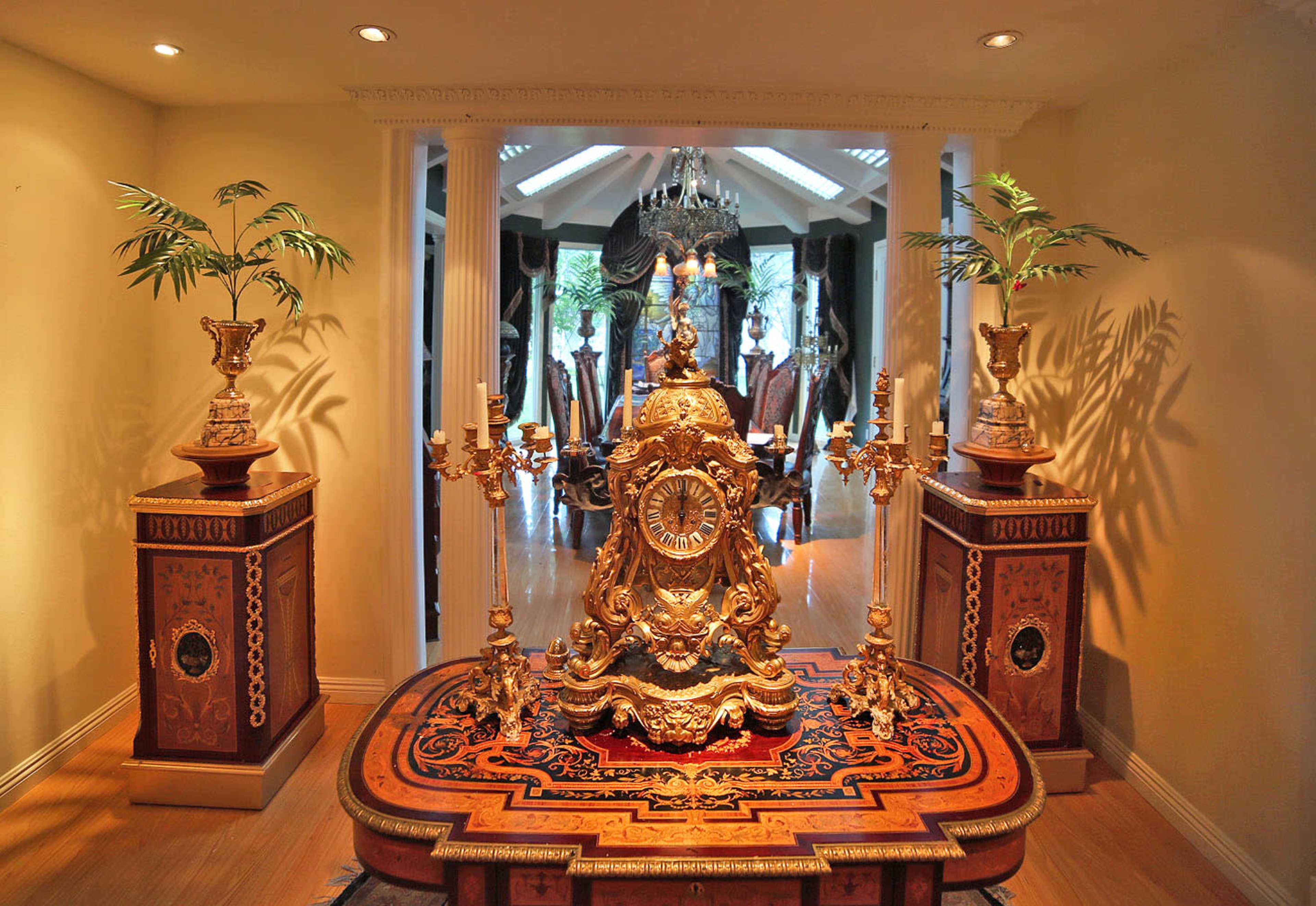 The image features an ornate clock centerpiece on a decorative table, flanked by two large planters, with a luxurious room visible in the background.