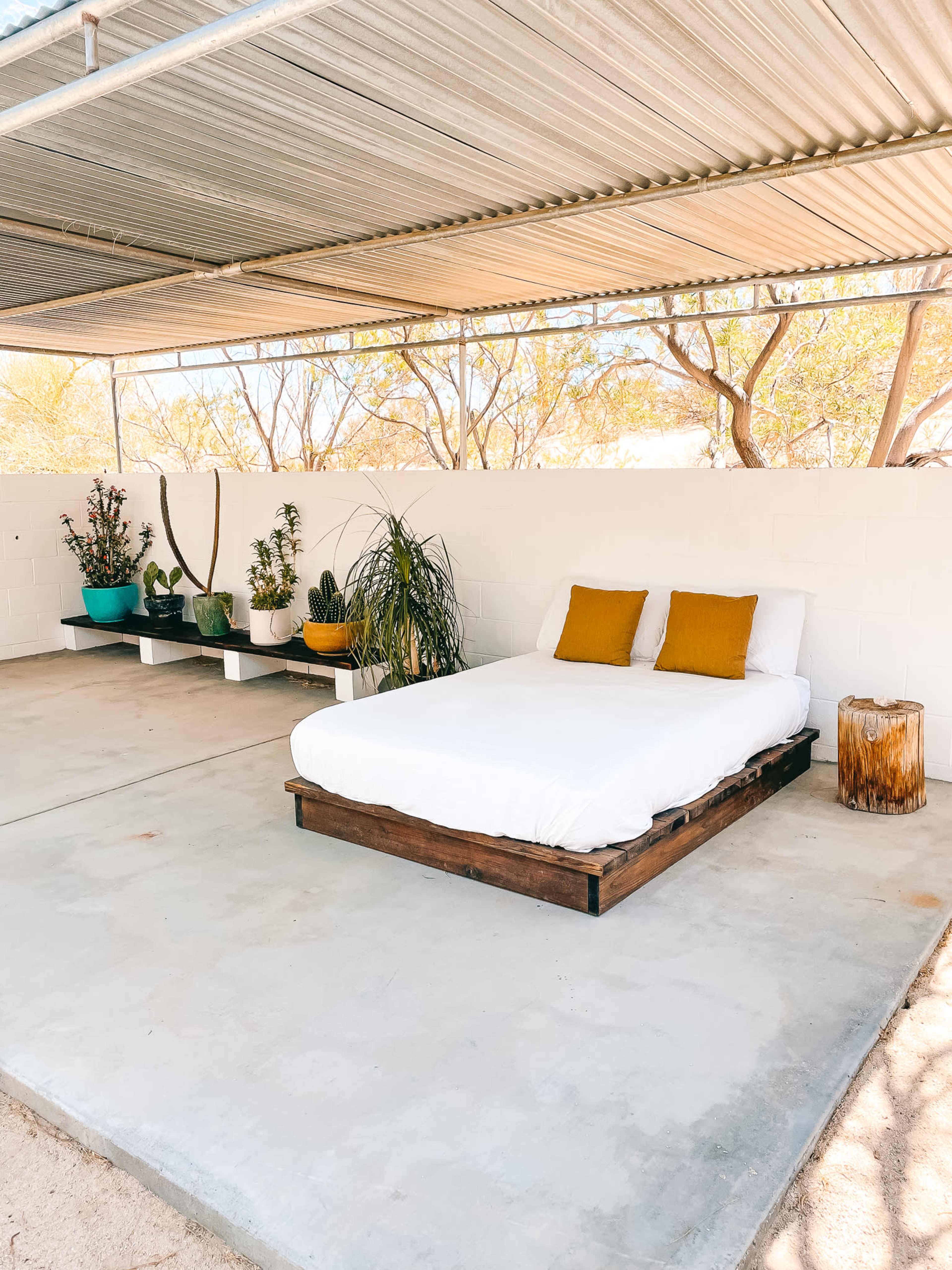 The image shows a minimalistic outdoor sleeping area with a white bed on a wooden frame, accompanied by potted plants and a small wooden stool.