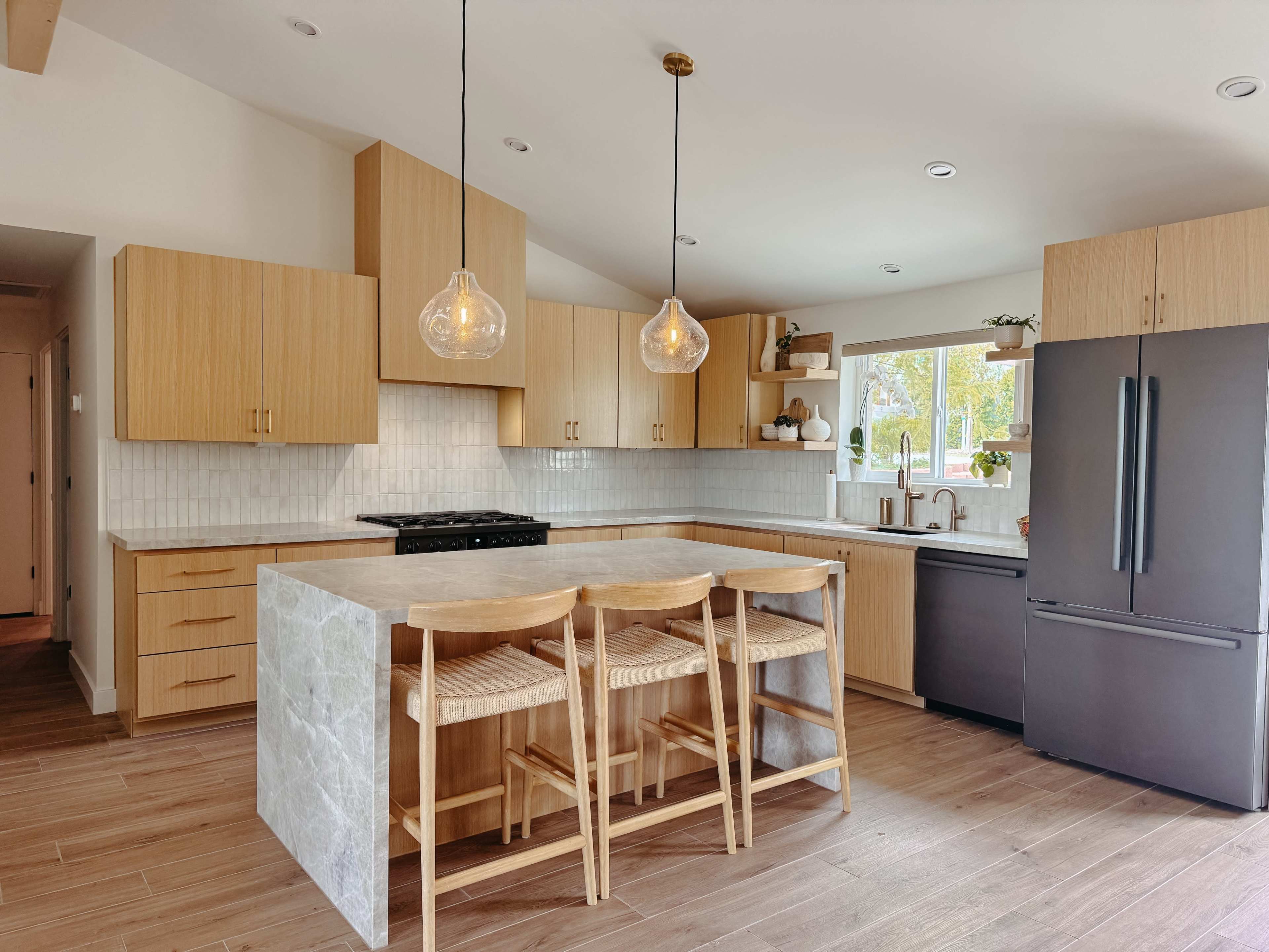 The image shows a modern kitchen with wooden cabinets, a large island featuring gray stone, and bar stools, illuminated by pendant lights.