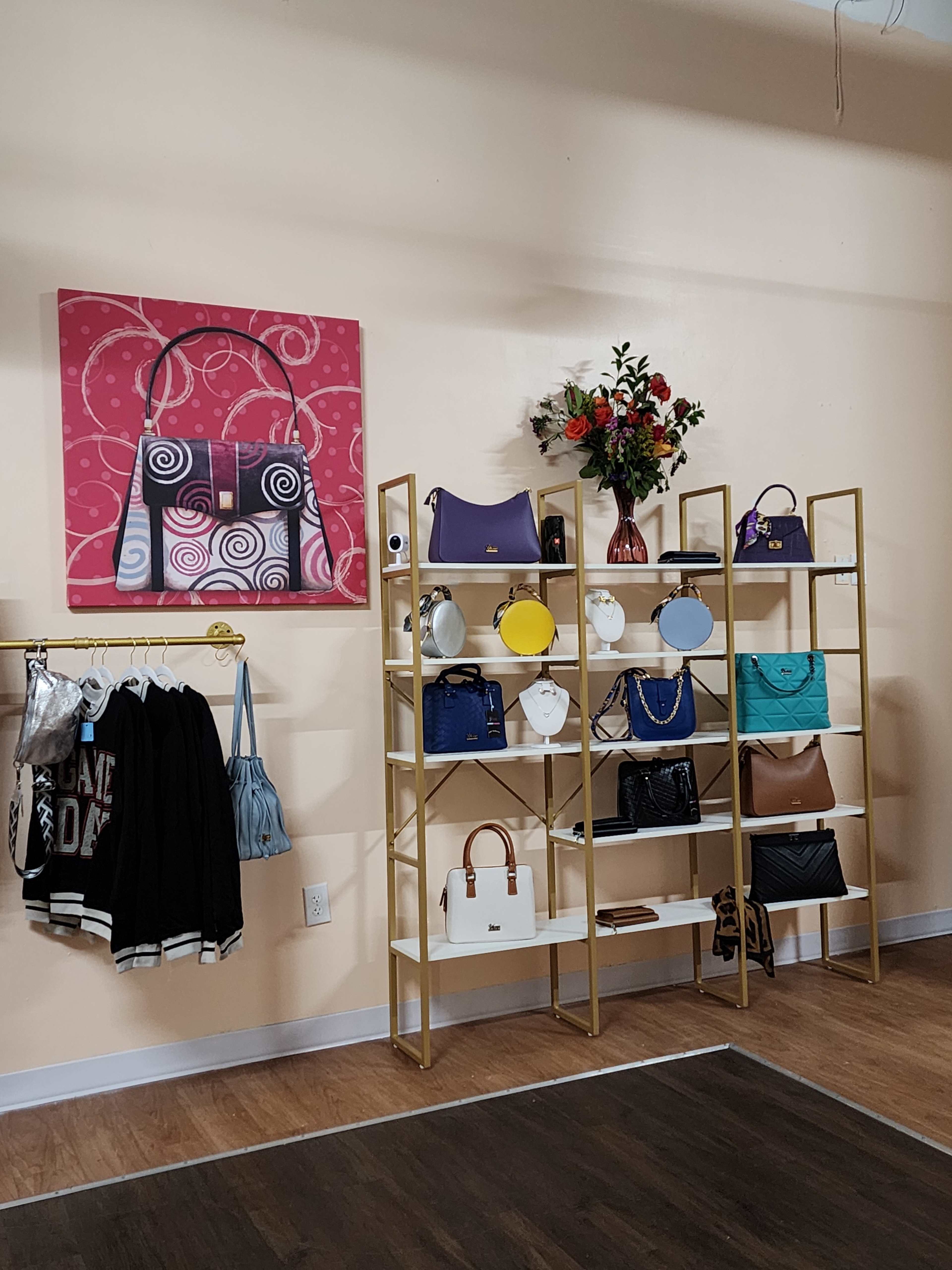 The image shows a boutique interior featuring a pink artwork on the wall, gold shelving displaying various handbags, and a floral arrangement on top of the shelves.