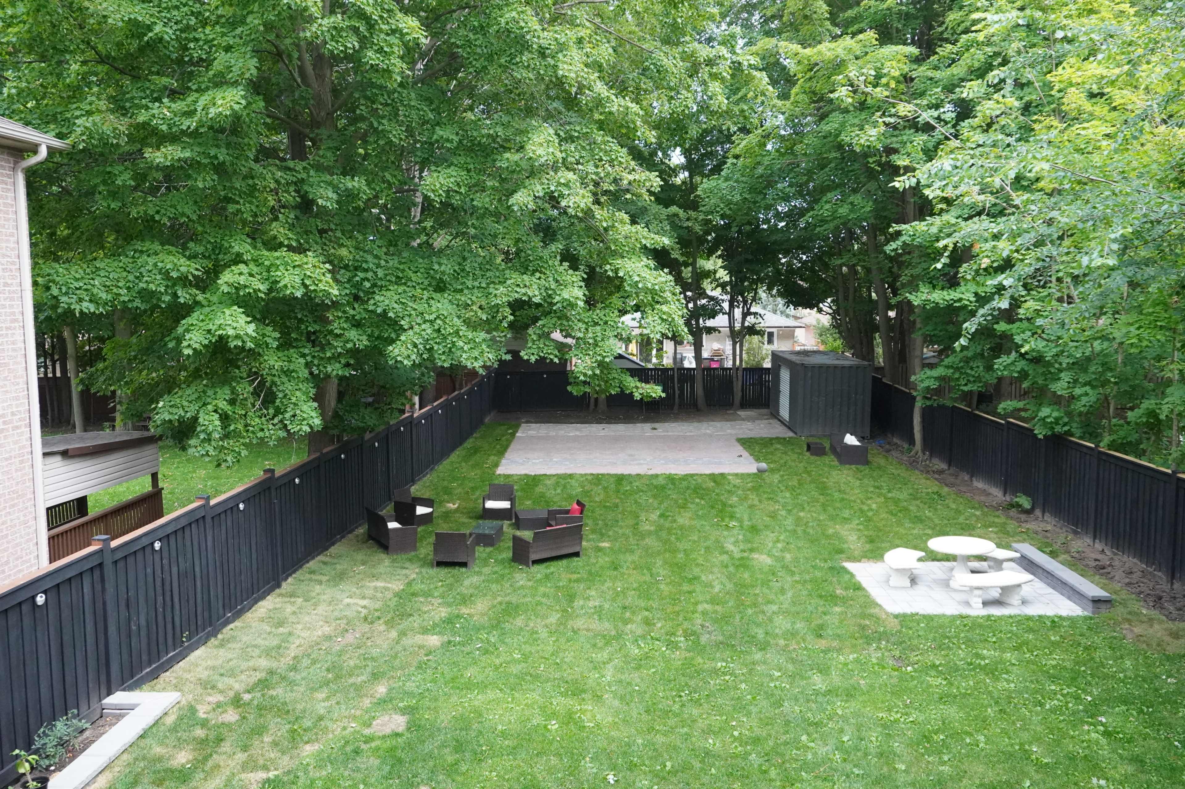 The image shows a spacious backyard with manicured grass, a large square patio area, a picnic table, and several outdoor chairs set against a backdrop of tall trees and a black wooden fence.