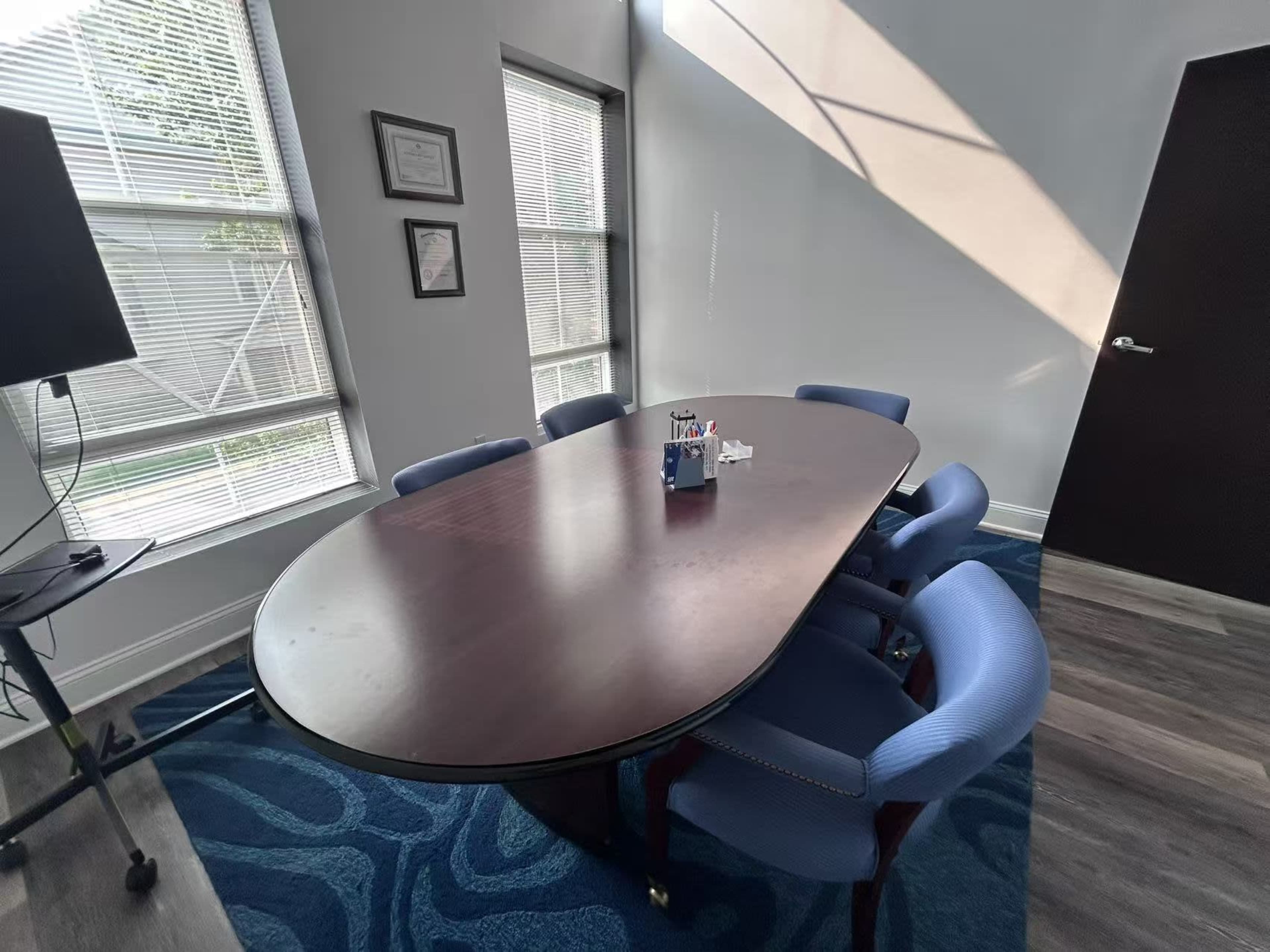 The image shows a conference room with a large oval table surrounded by five blue chairs, featuring windows that allow natural light to enter.