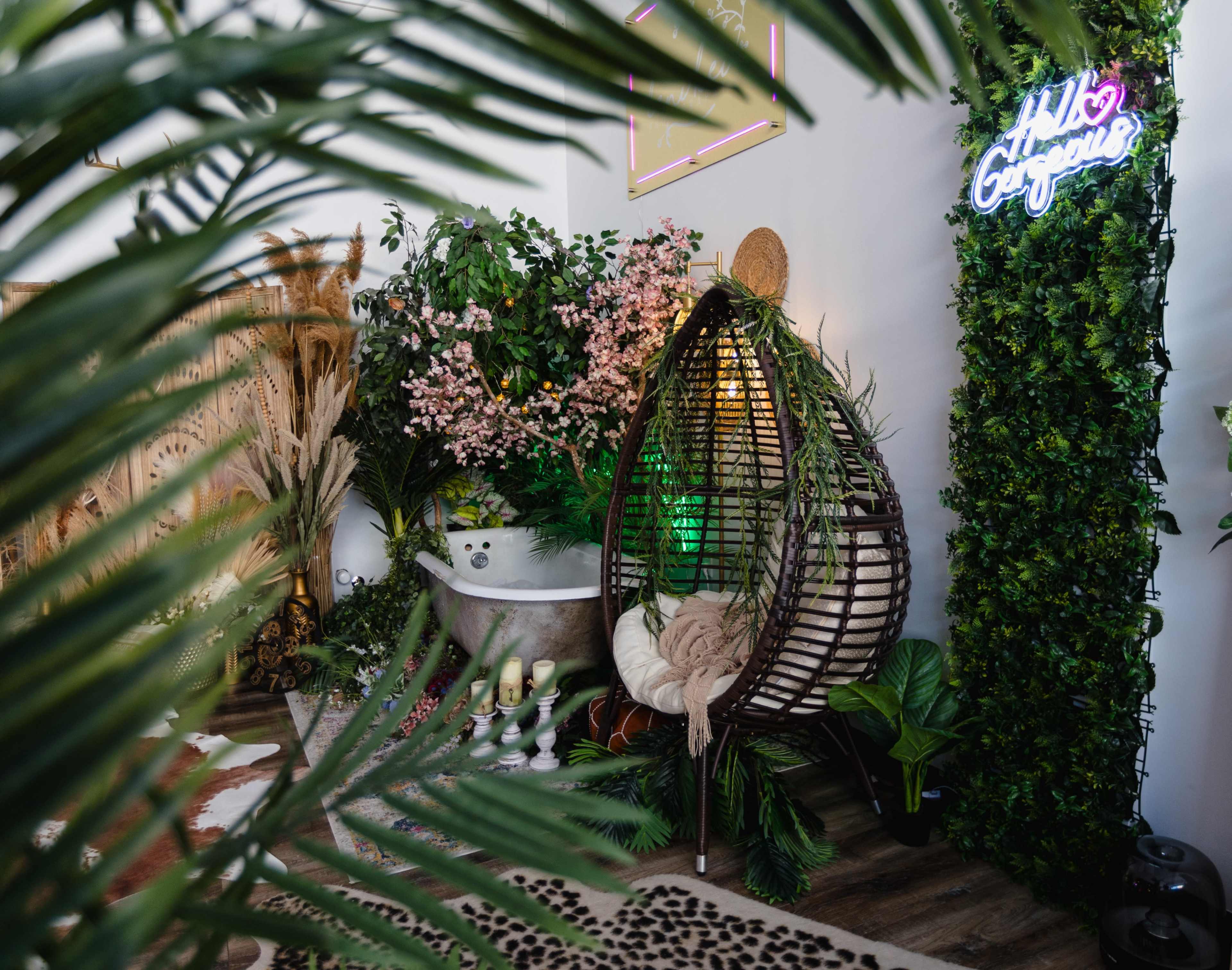 A cozy bathroom scene features a hanging wicker chair surrounded by lush greenery and floral arrangements, with a neon sign that says "Hello Gorgeous."