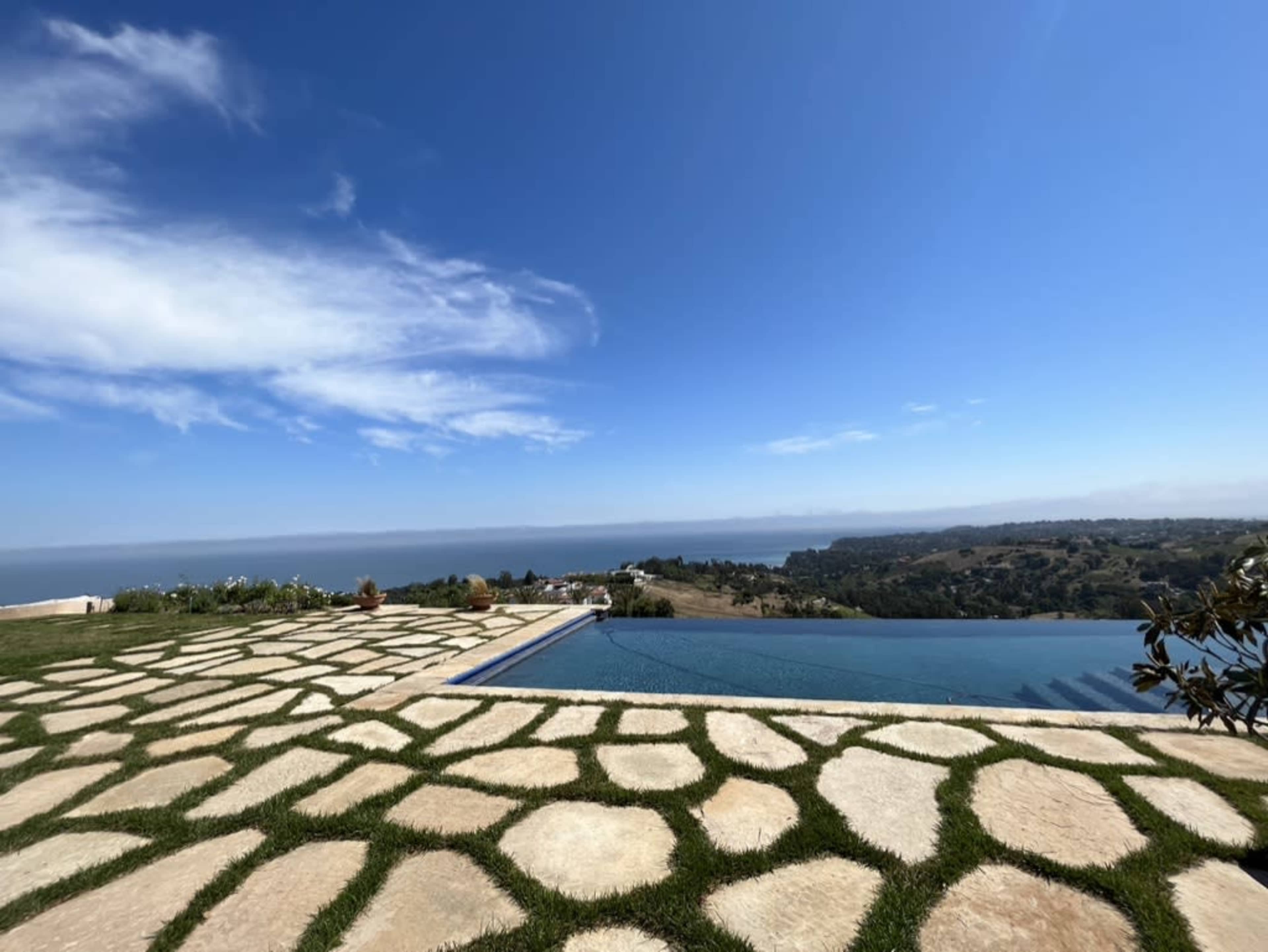 A stone patio with a swimming pool overlooking a scenic landscape and calm waters under a clear blue sky.