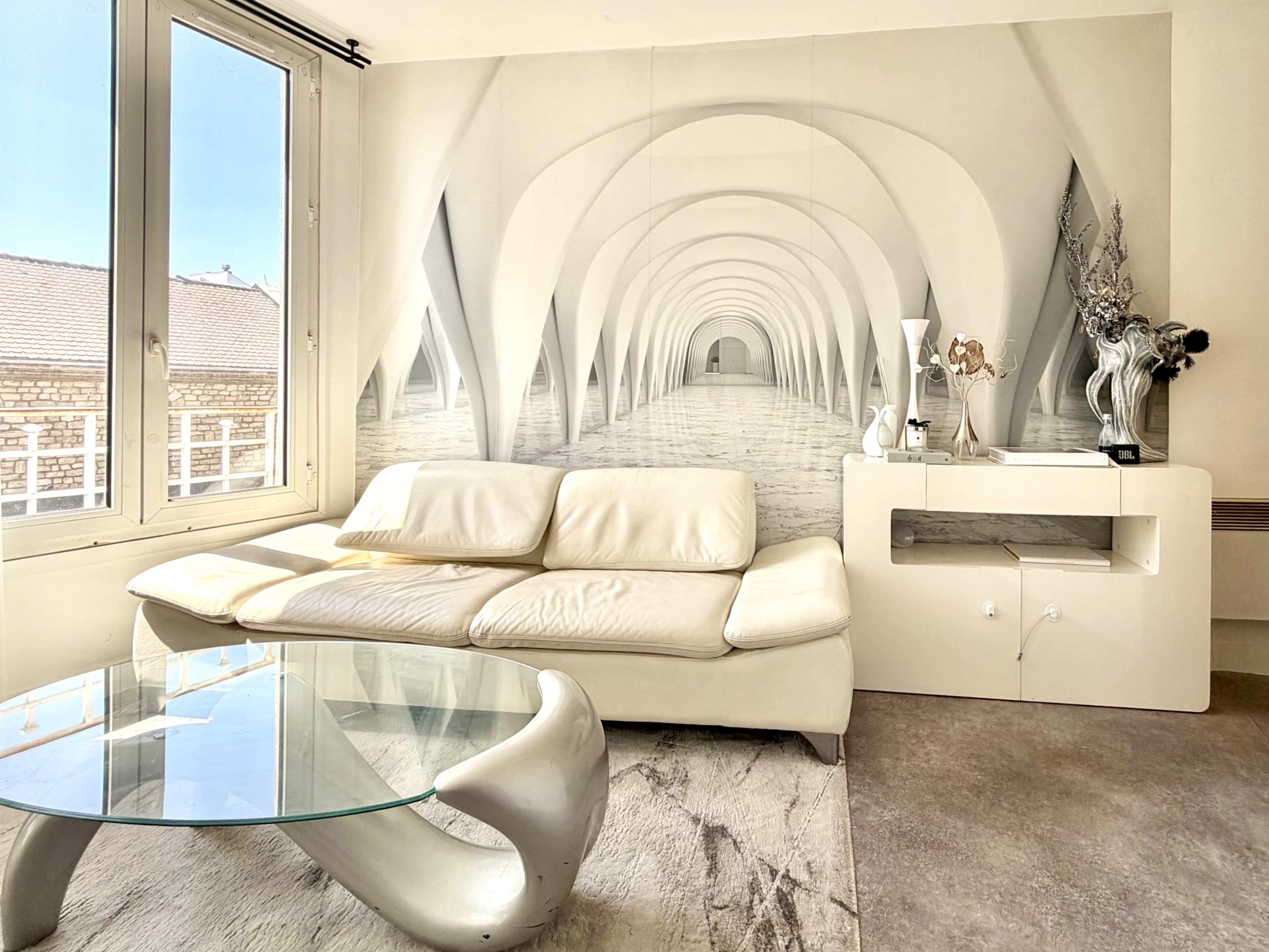 A modern living room features a white leather sofa, a glass coffee table, and a wall mural depicting an arched corridor.