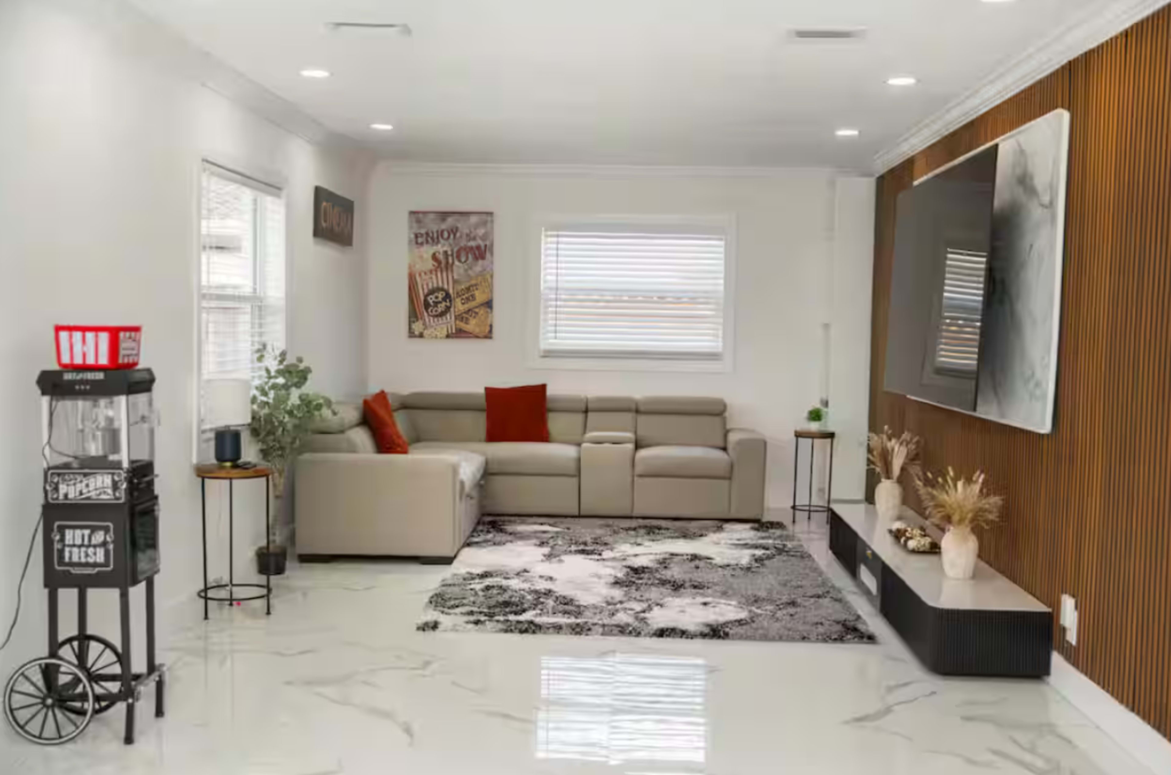 The image shows a modern living room featuring a beige sectional sofa, a large television mounted on a wooden accent wall, and a black and white area rug.