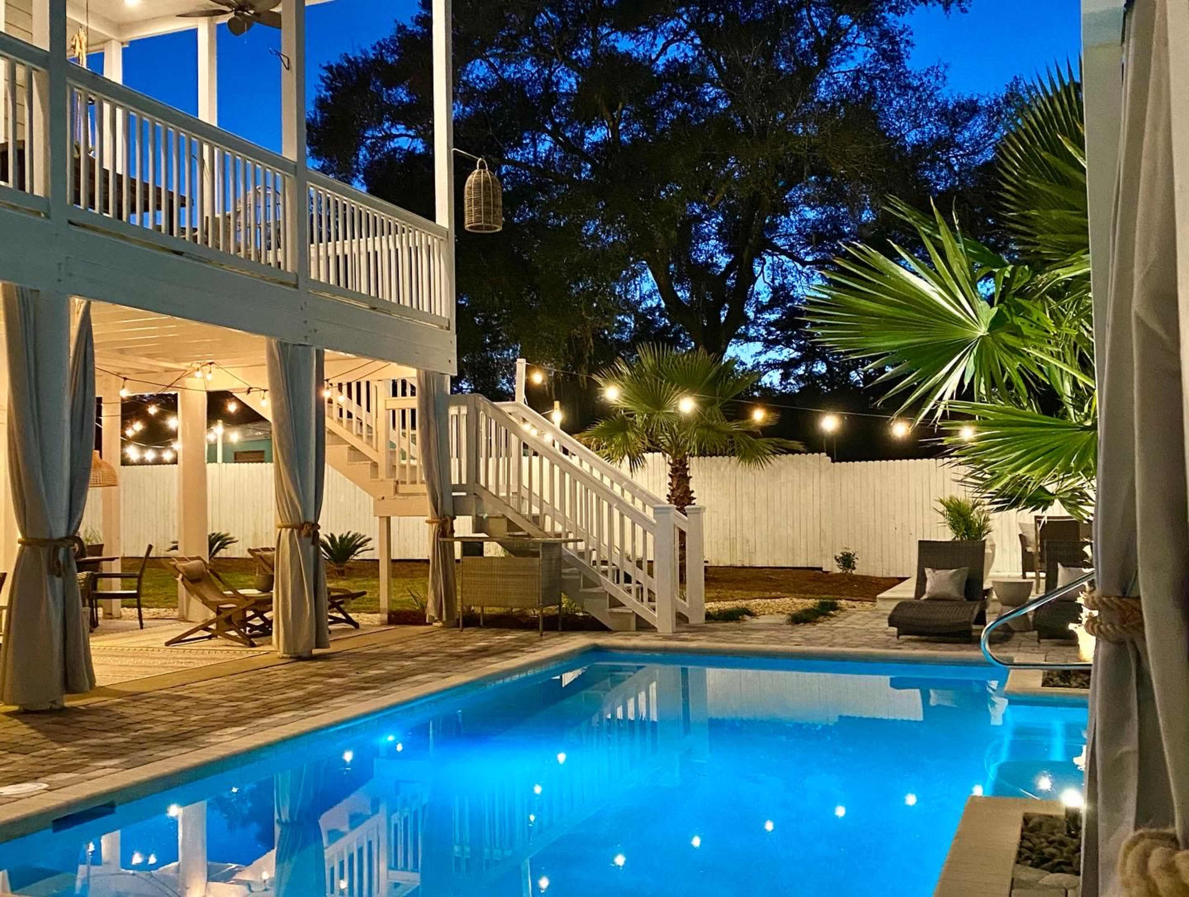 A well-lit outdoor pool area with a staircase leading to a raised deck, surrounded by palm trees and cozy seating.