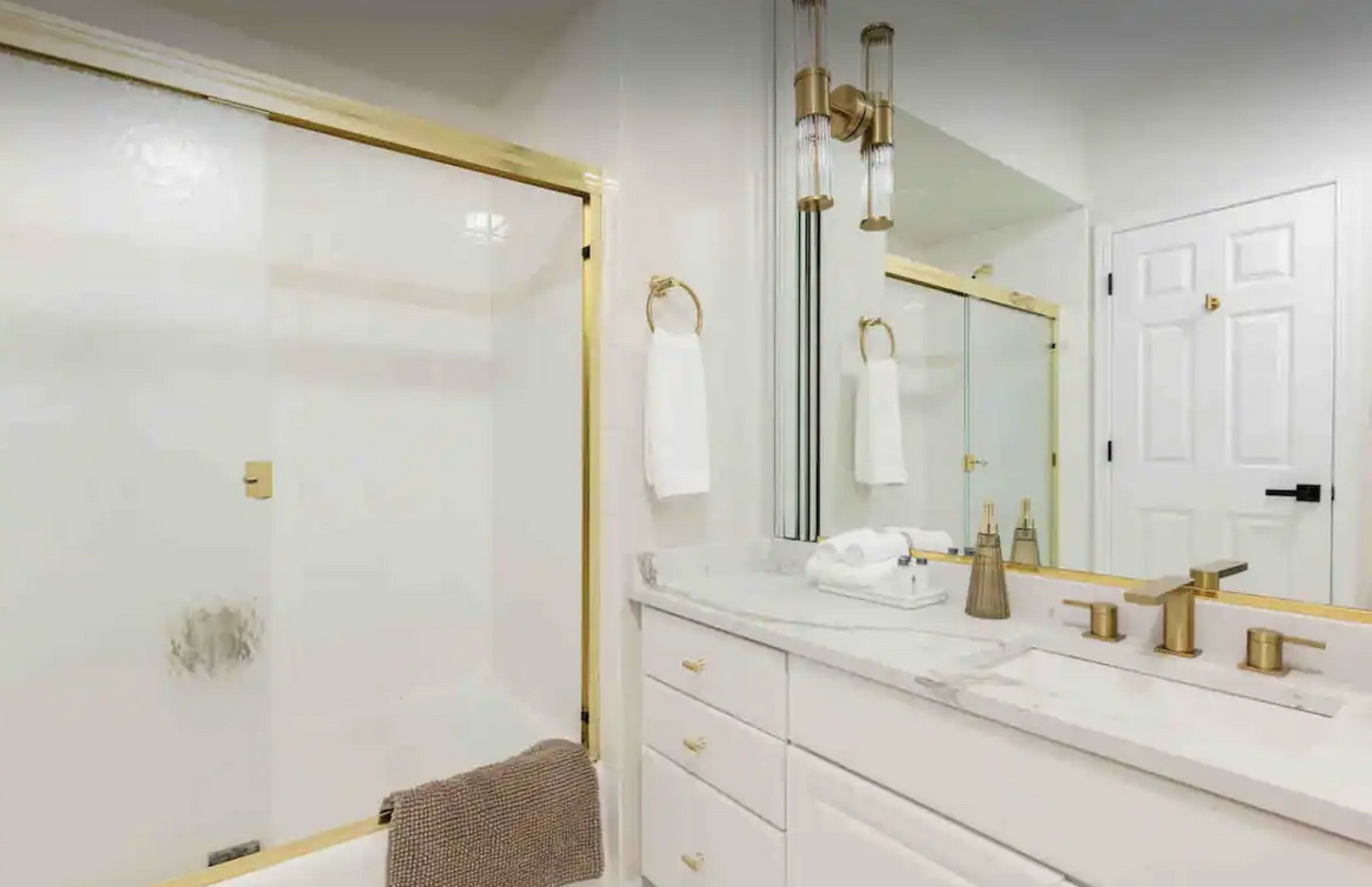 A bathroom with a glass shower door, double vanity with a marble countertop, and gold fixtures.