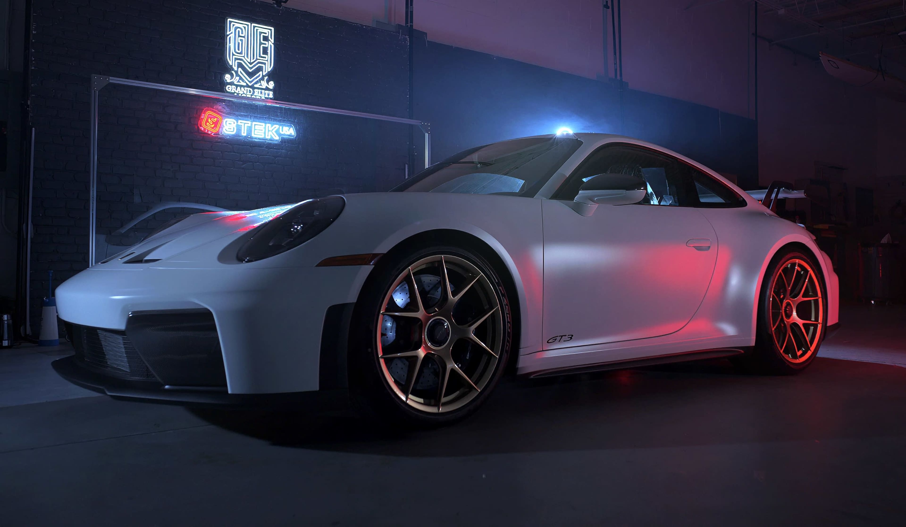 A white Porsche 911 GT3 is prominently displayed in a dimly lit garage, illuminated by colorful lights and a neon sign in the background.