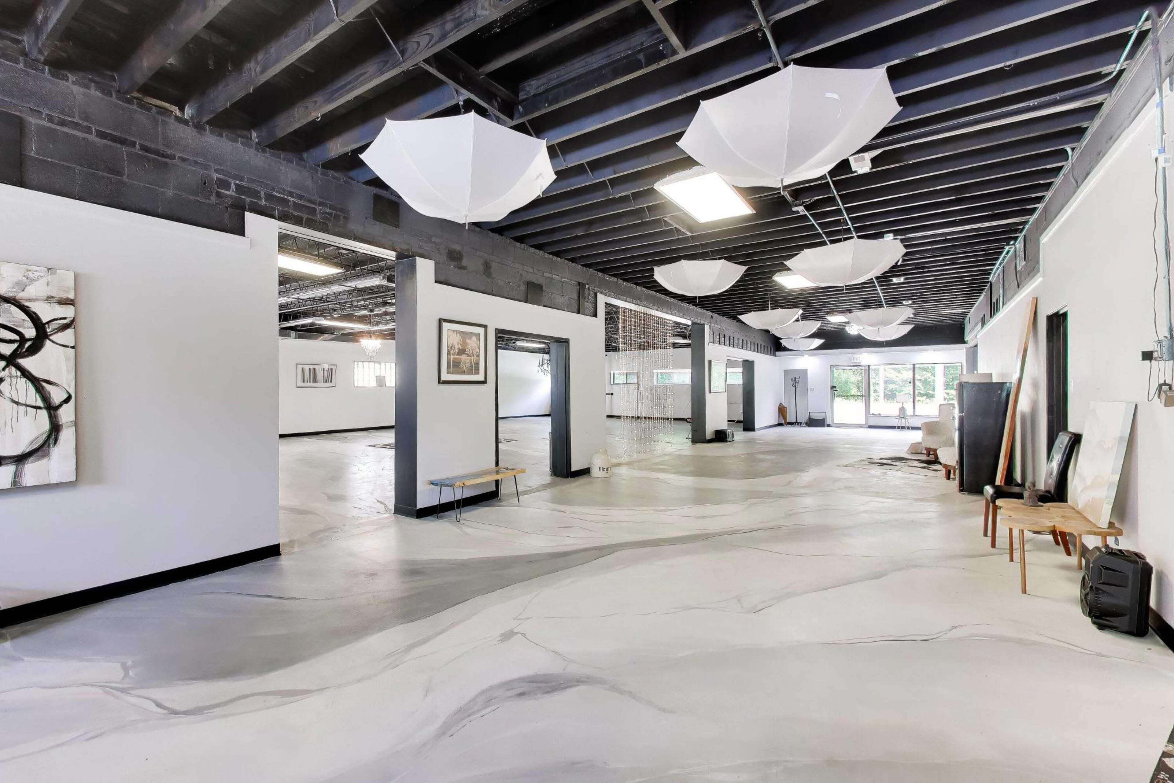 The image depicts a spacious, modern gallery with a polished concrete floor and white umbrellas hanging from the ceiling.