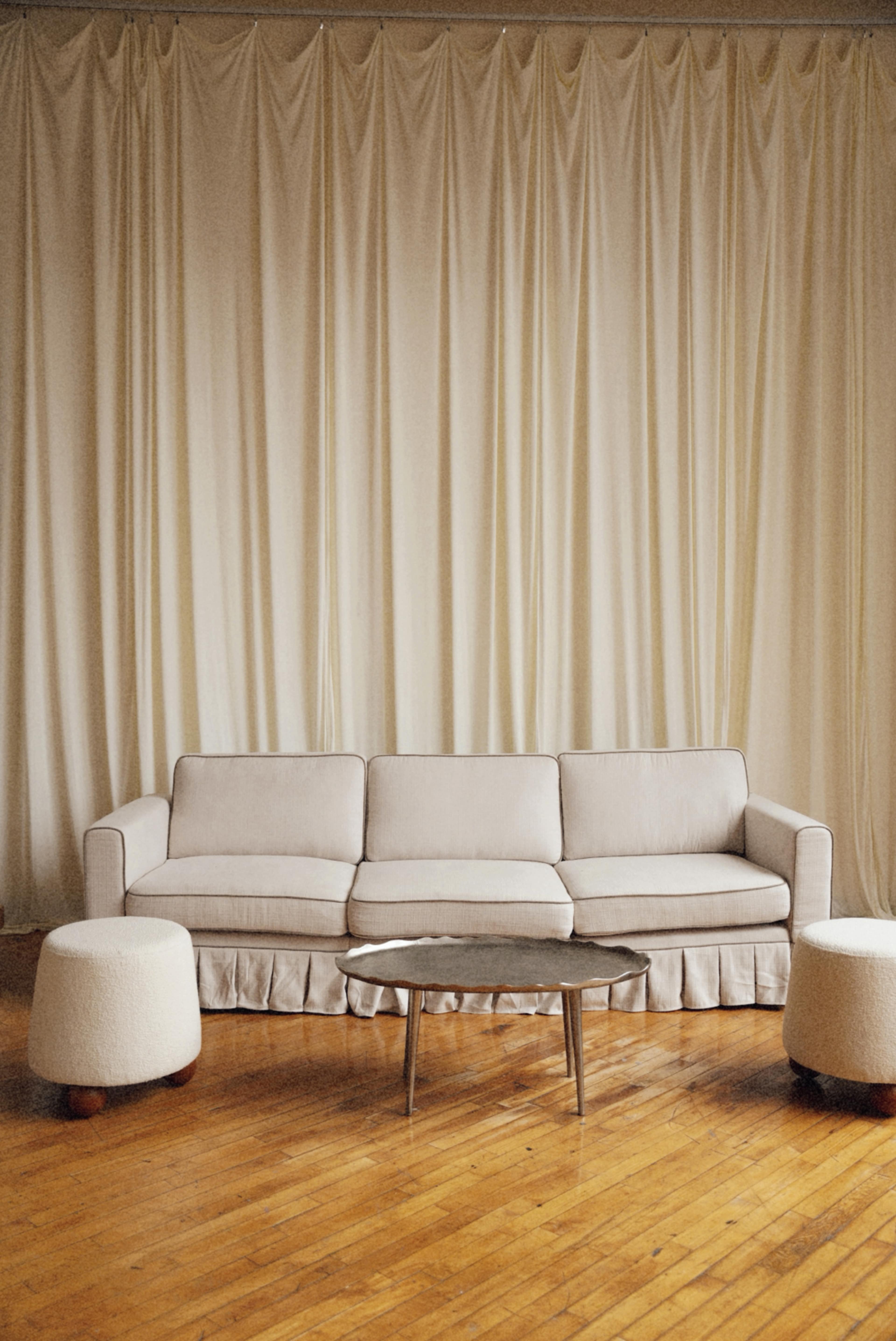 A light-colored sofa with a ruffled skirt is positioned in front of a wooden floor, accompanied by two round ottomans and a low coffee table, all against a backdrop of cream-colored drapes.
