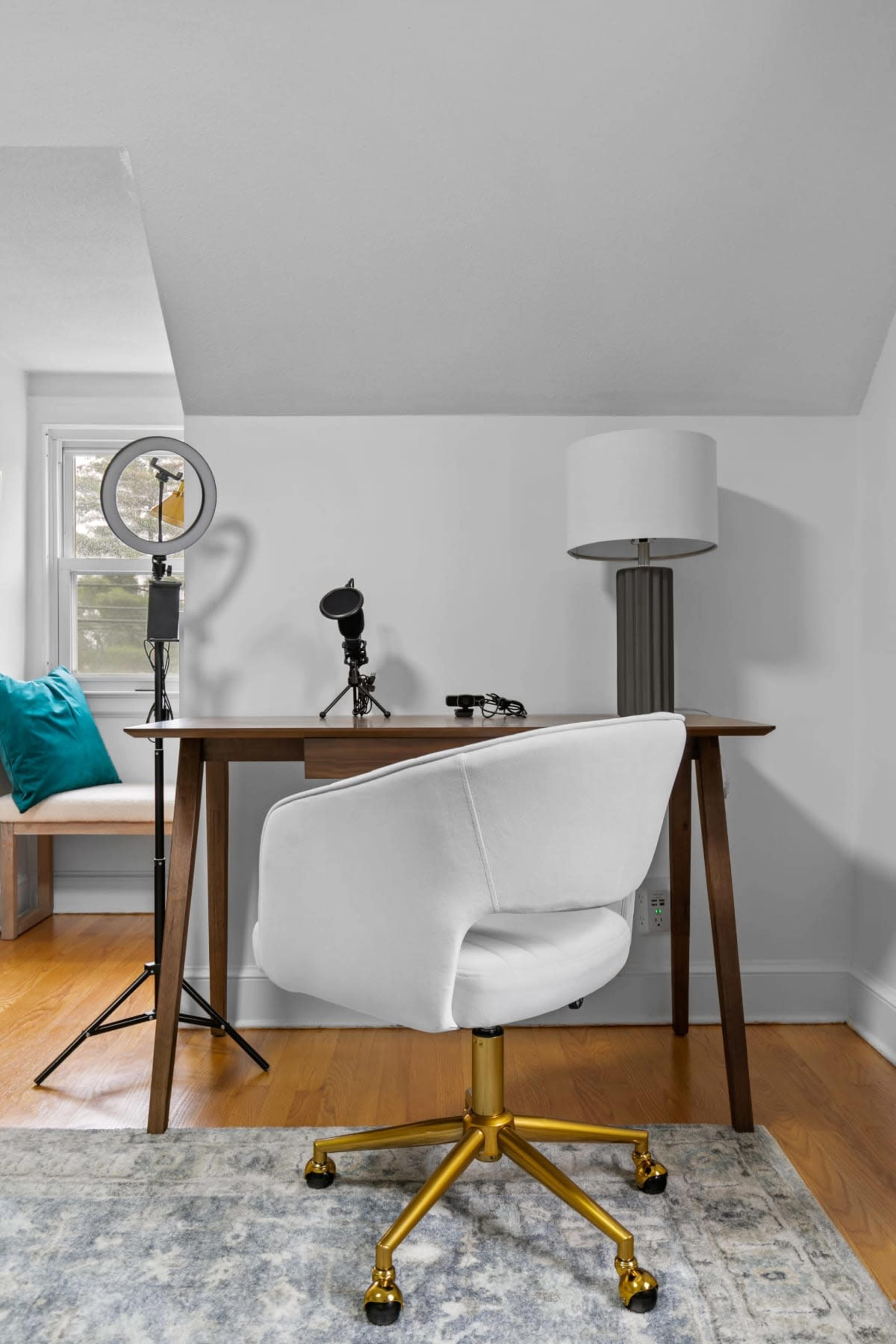 A simple desk with a white chair and a lamp is positioned in a well-lit room, accompanied by a ring light and microphones on stands.