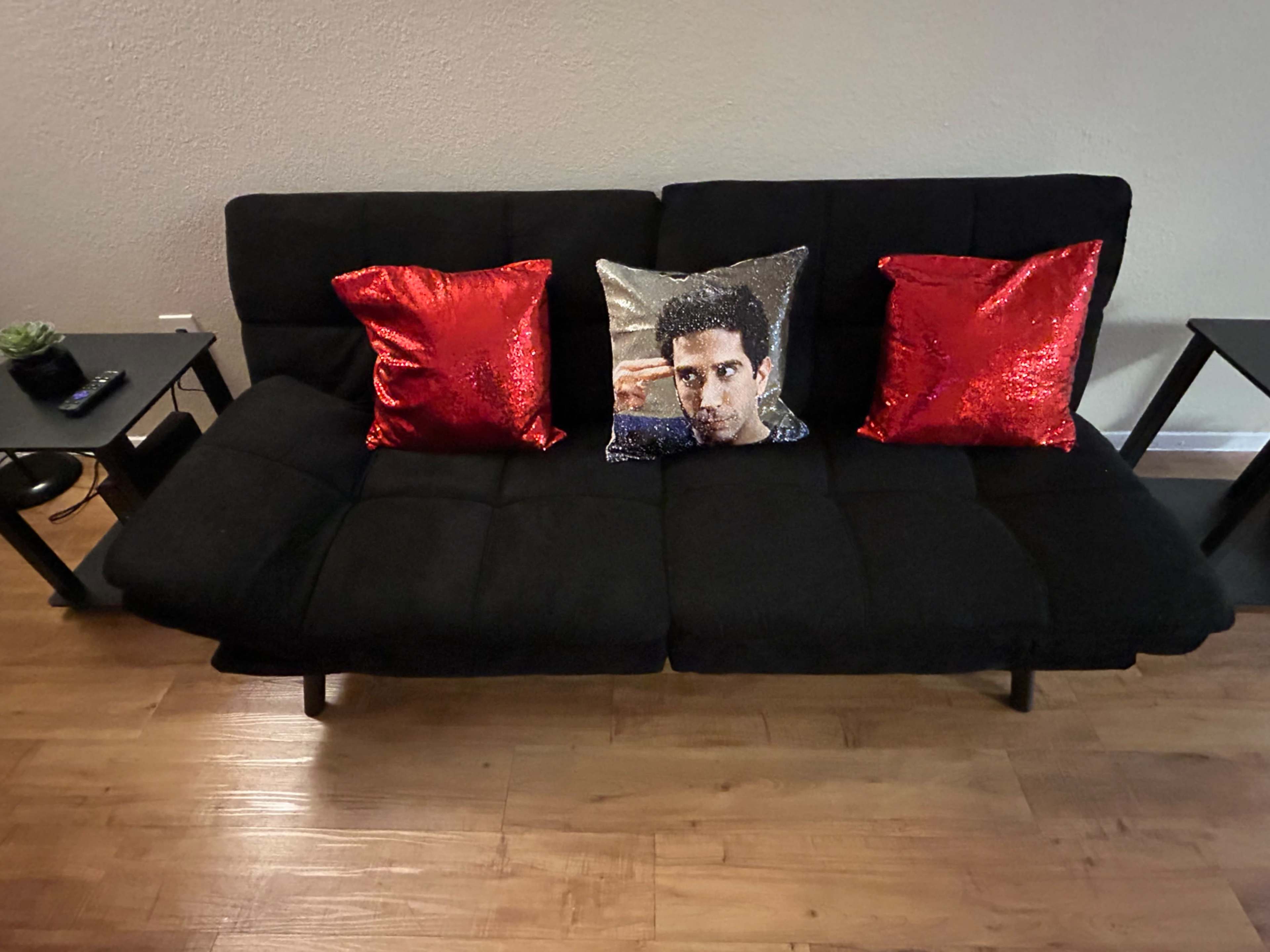 A black futon with two red sequined throw pillows and a decorative pillow featuring a man's image is positioned on a wooden floor.