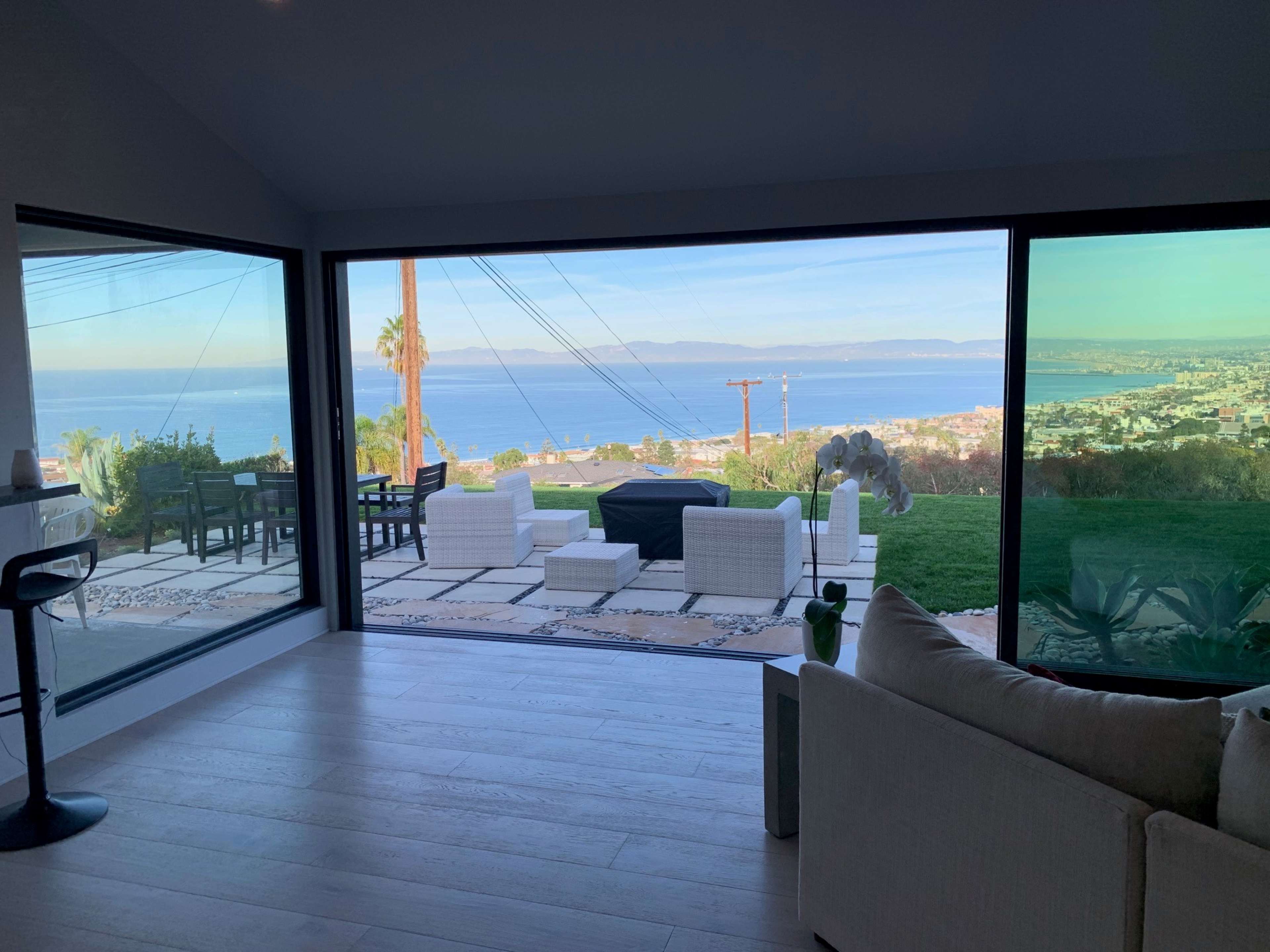 The image shows a modern interior space with large windows revealing a view of a coastal landscape, including a body of water and nearby city.