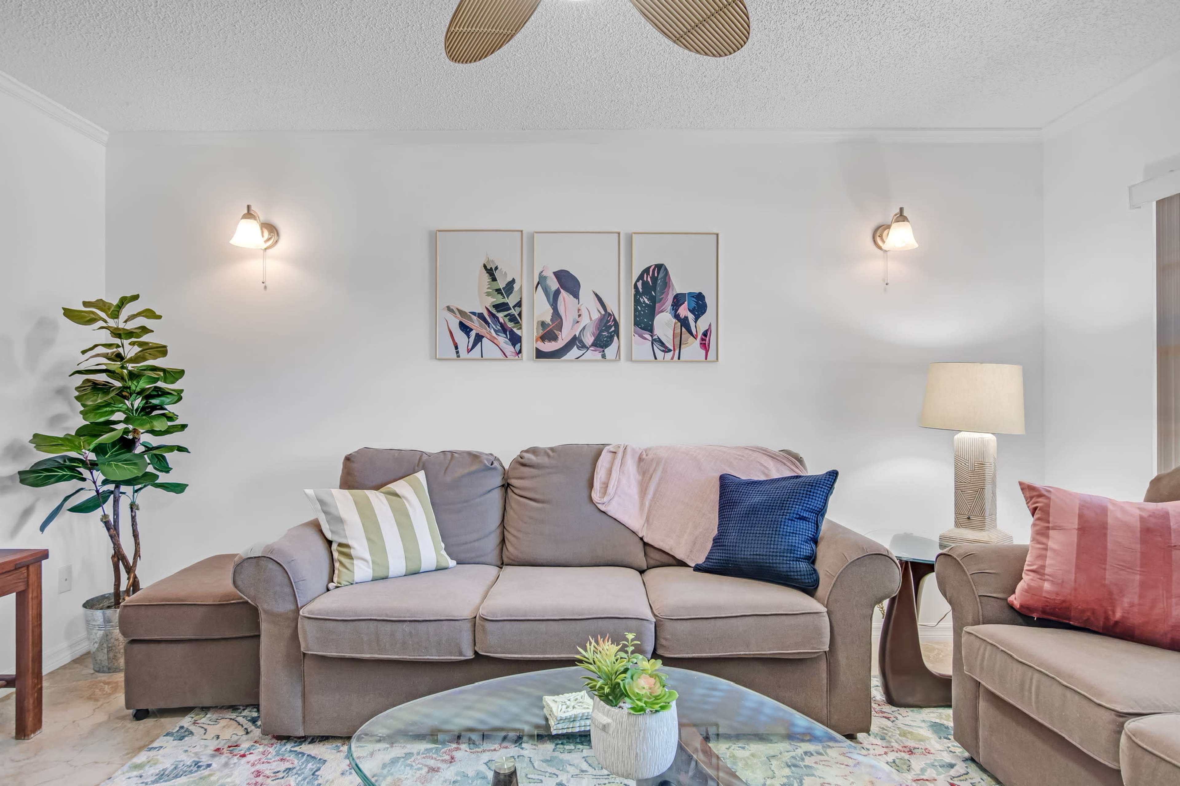 A cozy living room features a beige sofa with decorative cushions, a glass coffee table, and framed leaf prints on the wall, accompanied by a floor lamp and a potted plant.