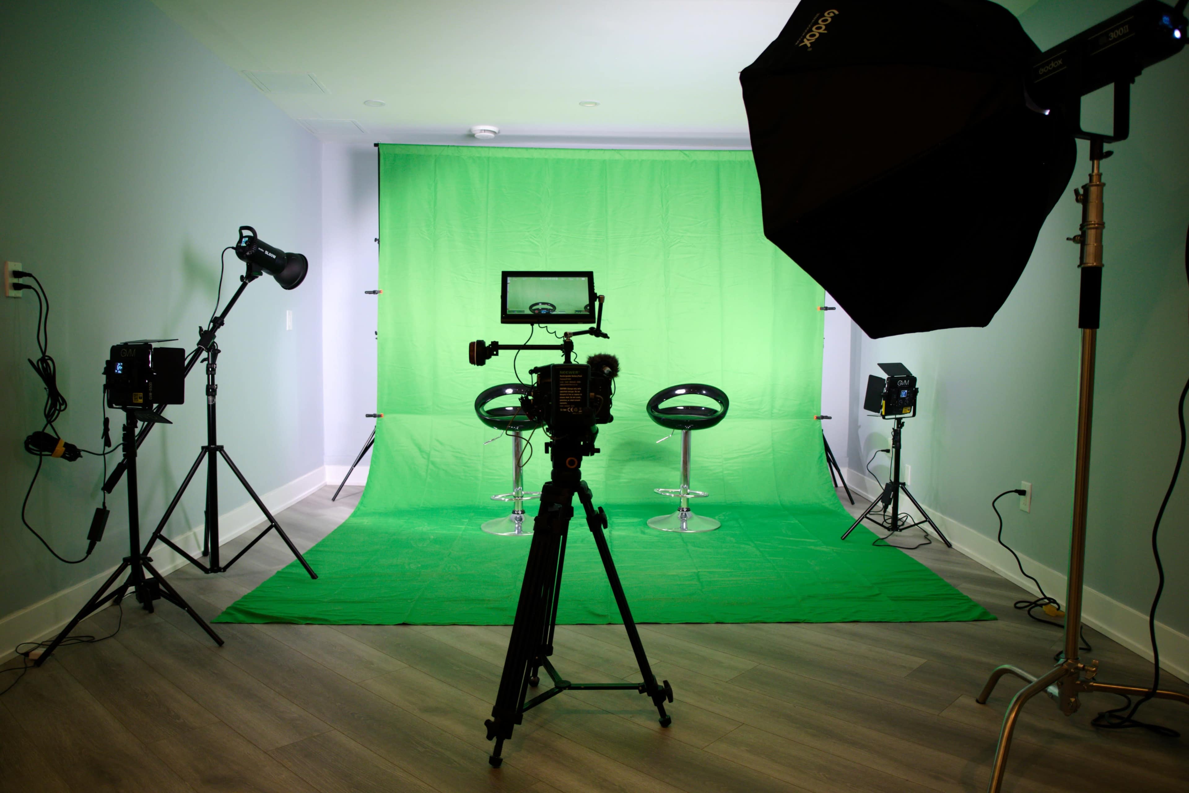 A video production studio is set up with a green backdrop, two bar stools, and various lighting equipment.