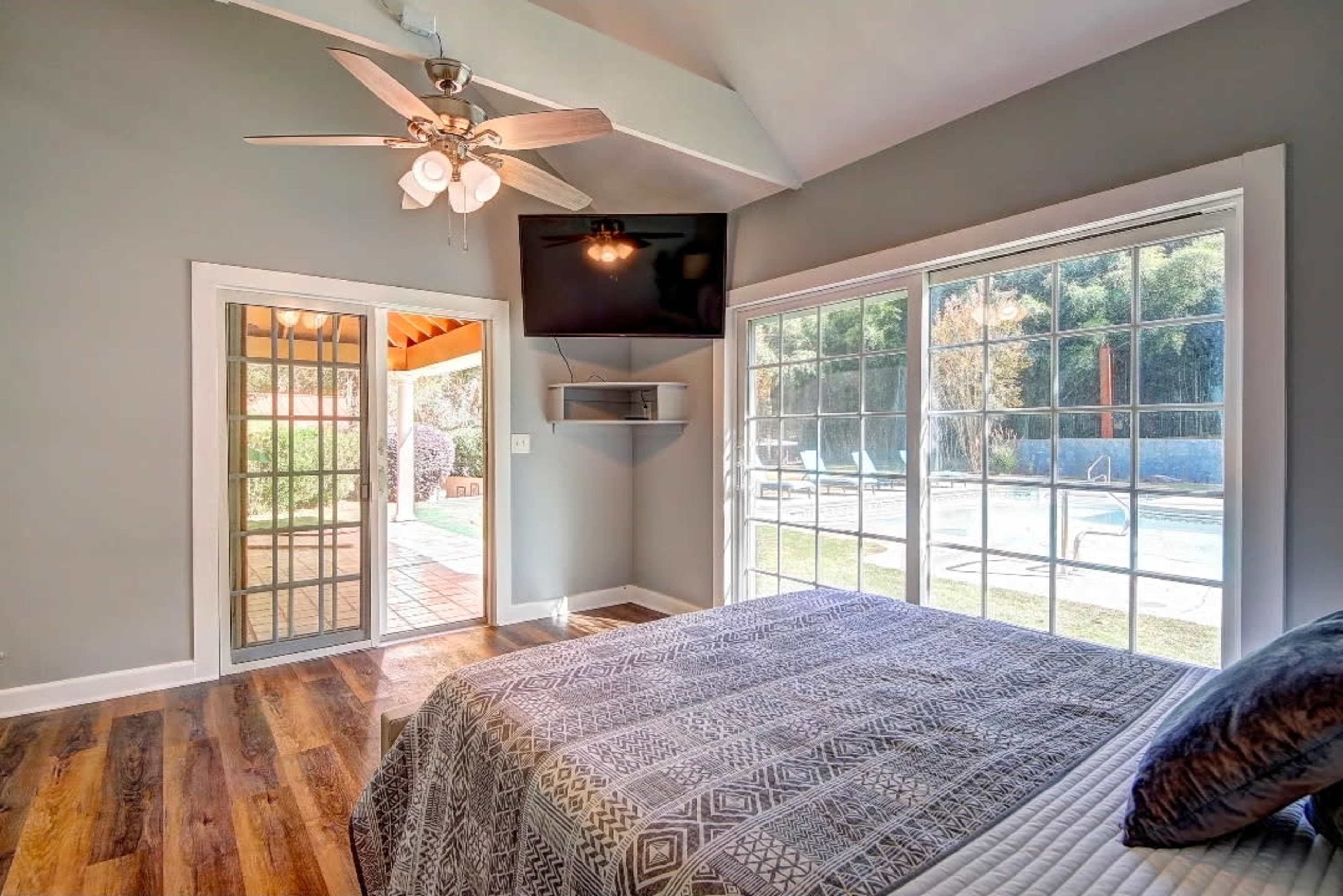 The image shows a bedroom with a patterned bedspread, large windows facing a pool area, and a ceiling fan.