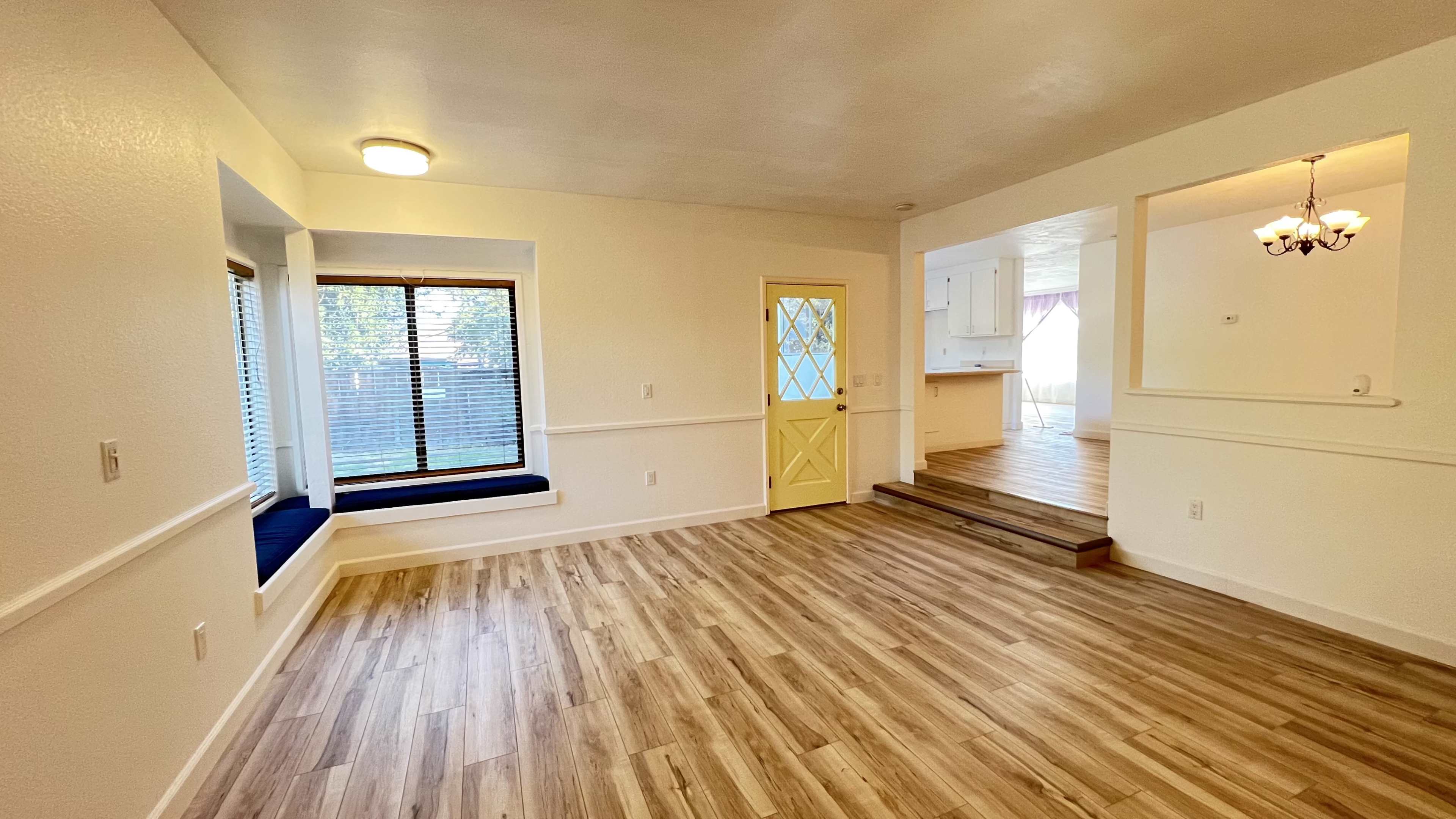 The image shows a light-filled interior space featuring a window seat, yellow door, and an open layout connecting to a kitchen area.