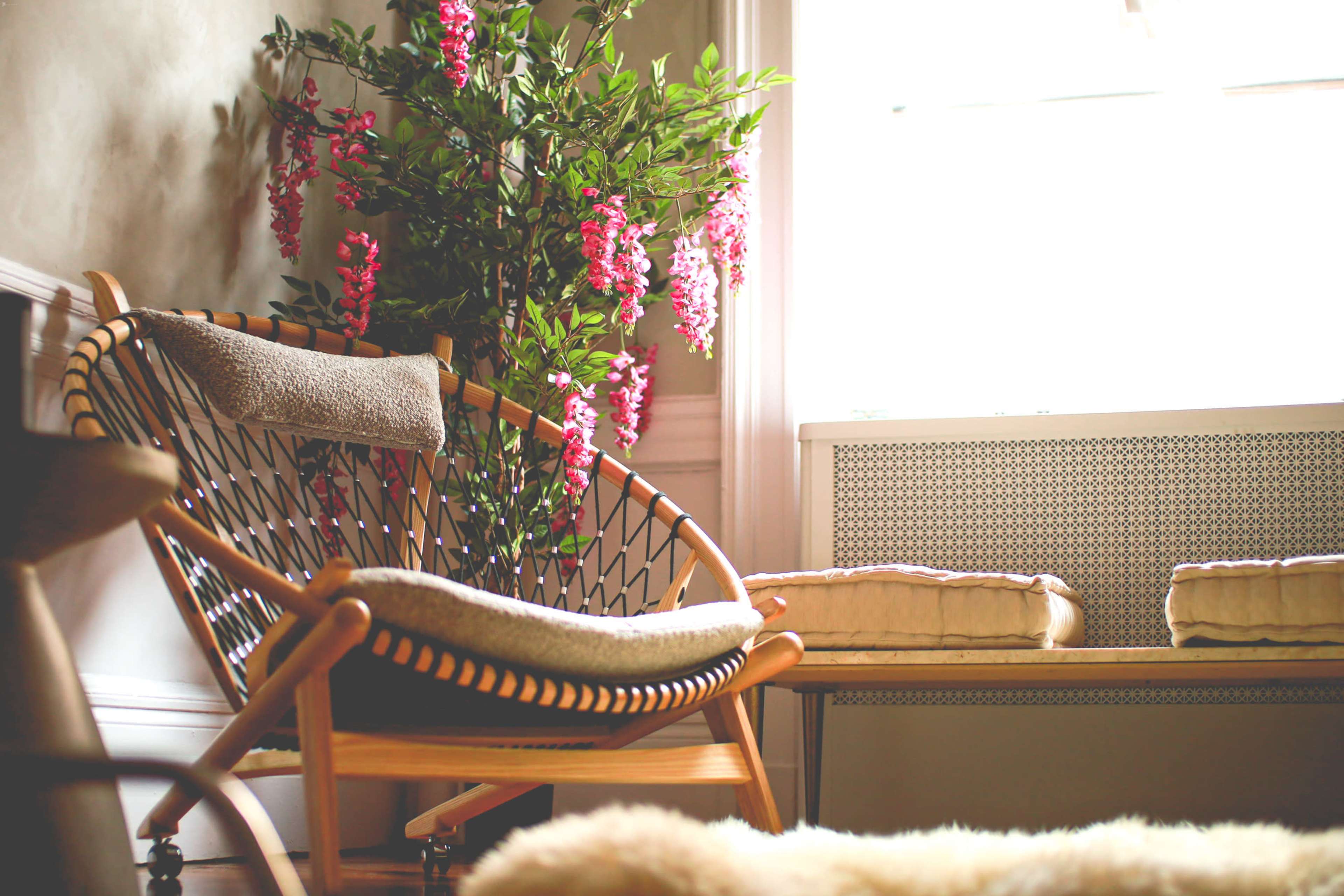 A wooden chair with a woven backrest sits near a window, accompanied by a plant with pink flowers and a bench with cushions.