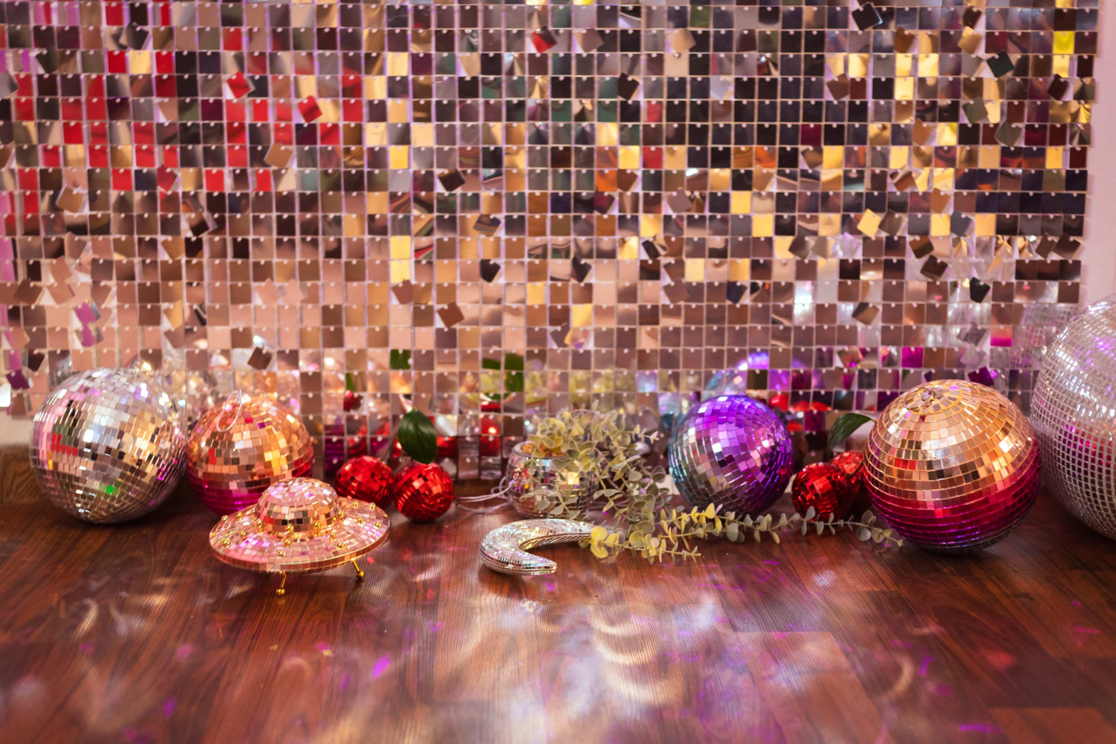 The scene features a sparkling backdrop of mirrored panels with various colorful disco balls, decorative ornaments, and greenery scattered on a wooden floor.
