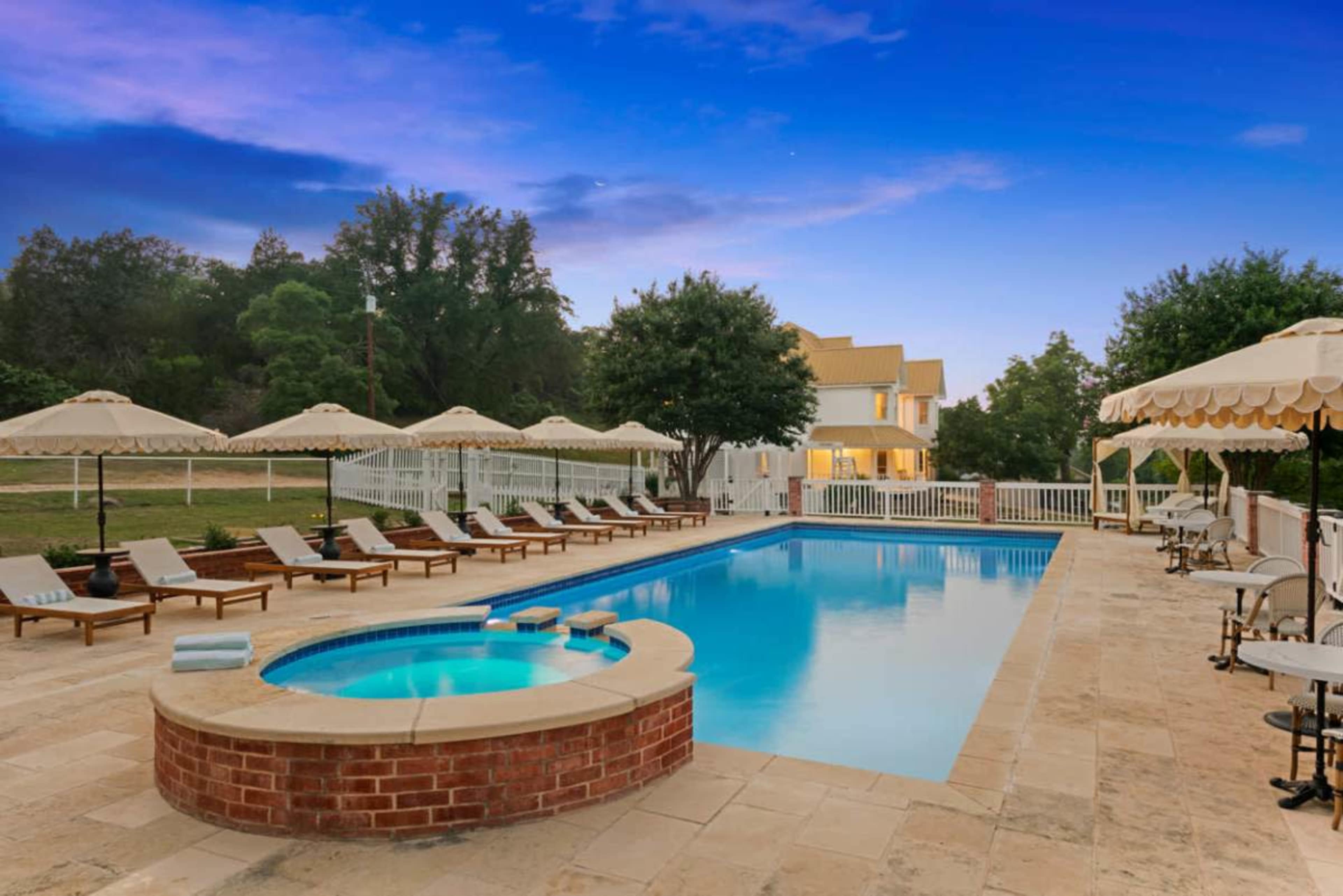 The image shows a swimming pool surrounded by lounge chairs and umbrellas, with a house visible in the background under a twilight sky.