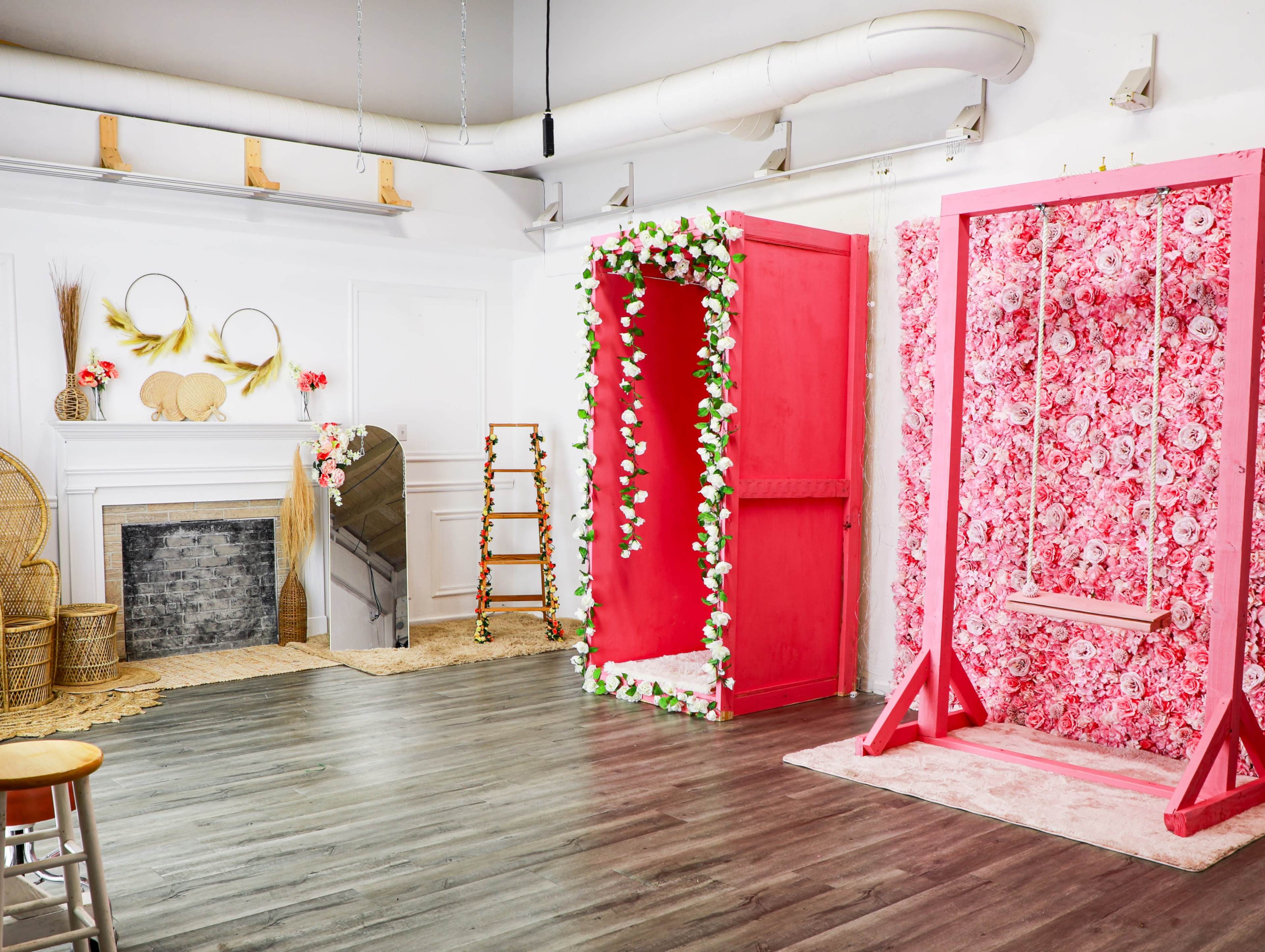 The image shows a bright, decorated studio space with a large pink floral backdrop, a flower-adorned wooden frame, and a swing, all arranged around a neutral-colored fireplace.
