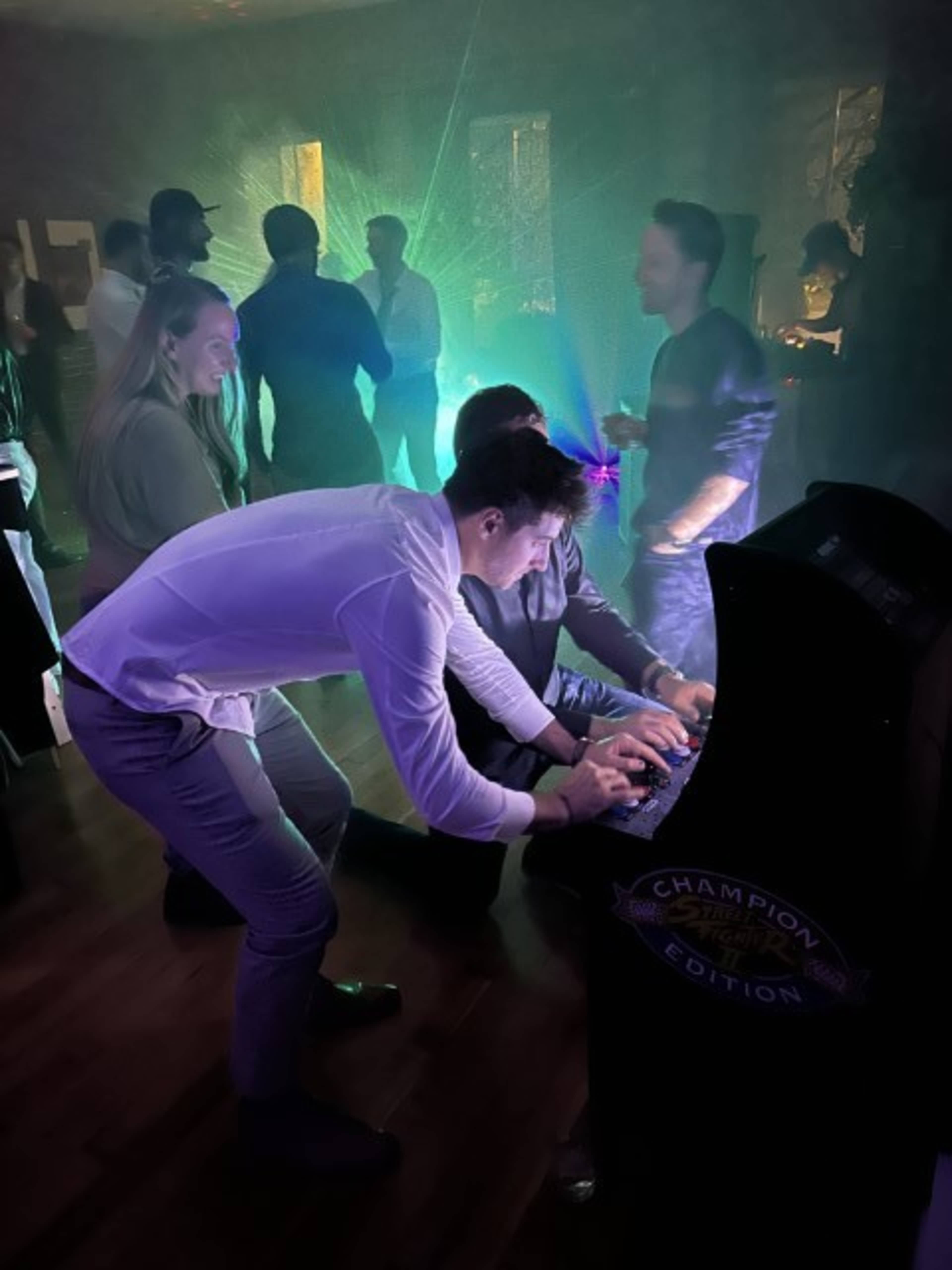 A group of people interacts around an arcade game in a dimly lit room with colorful lights.