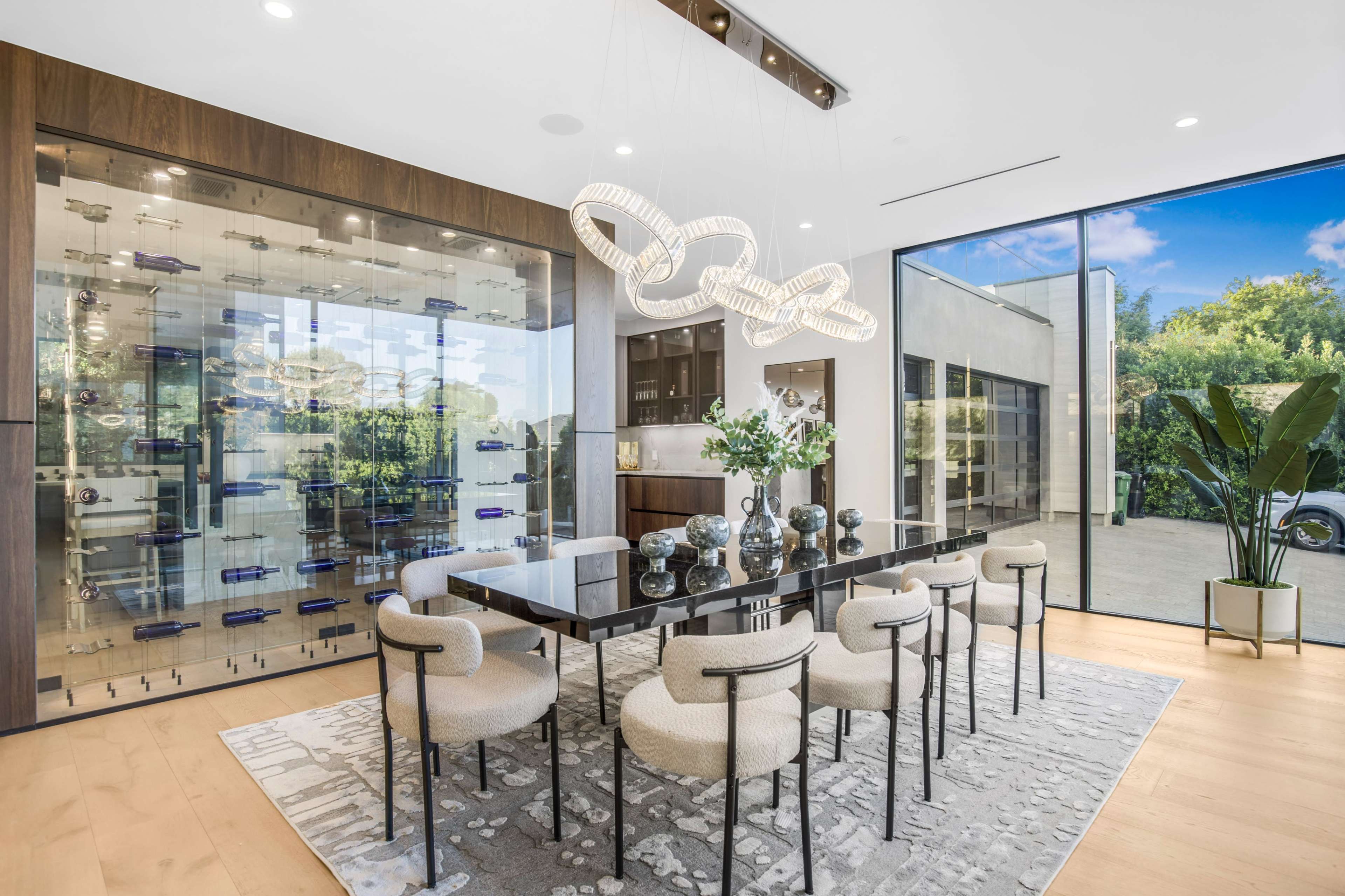A modern dining room features a large black table surrounded by upholstered chairs, alongside a glass wine display and large windows offering a view of the outdoor area.