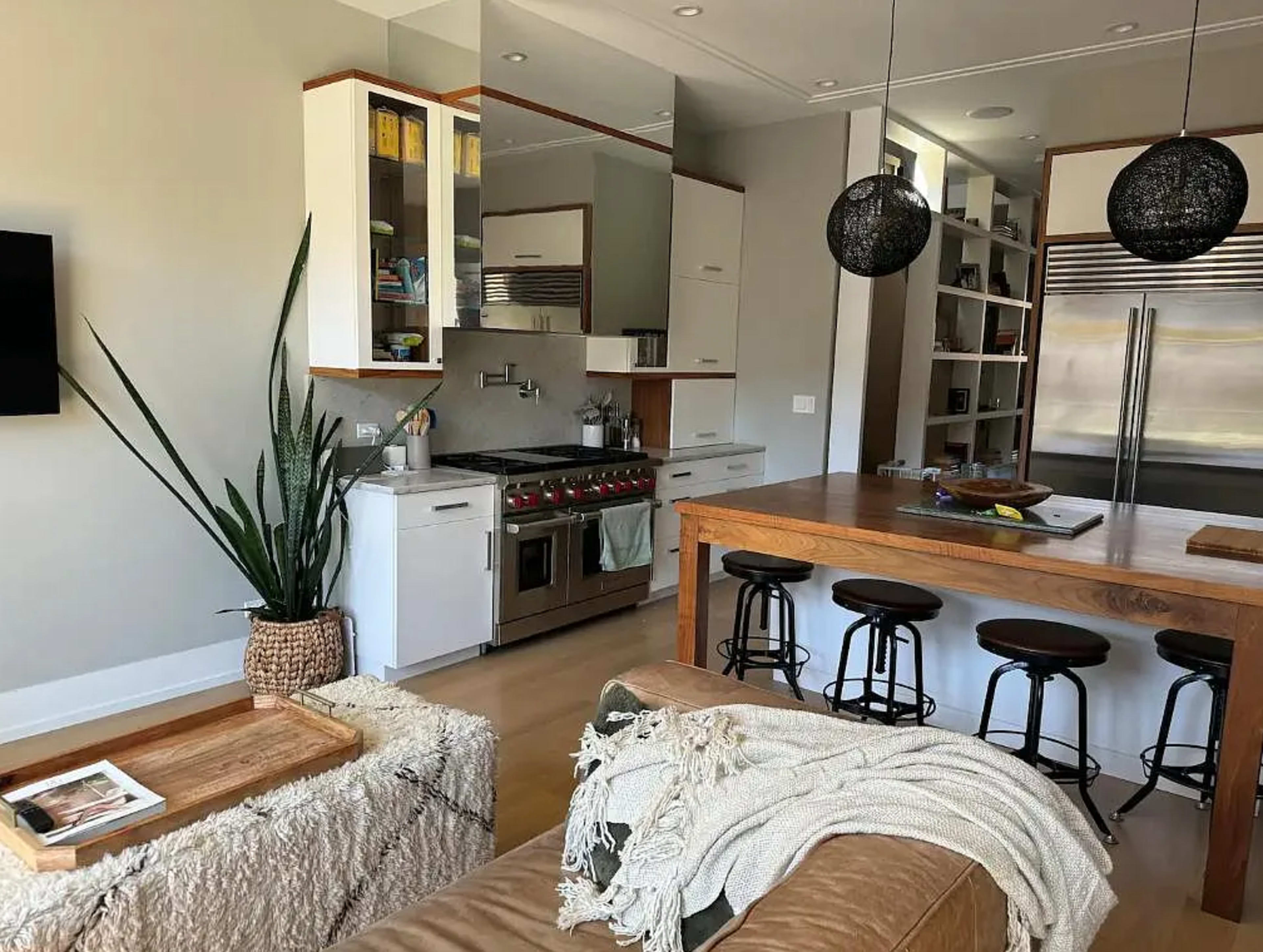 The image shows a modern kitchen and dining area featuring a wooden table with bar stools, stainless steel appliances, and a plant in a decorative pot.