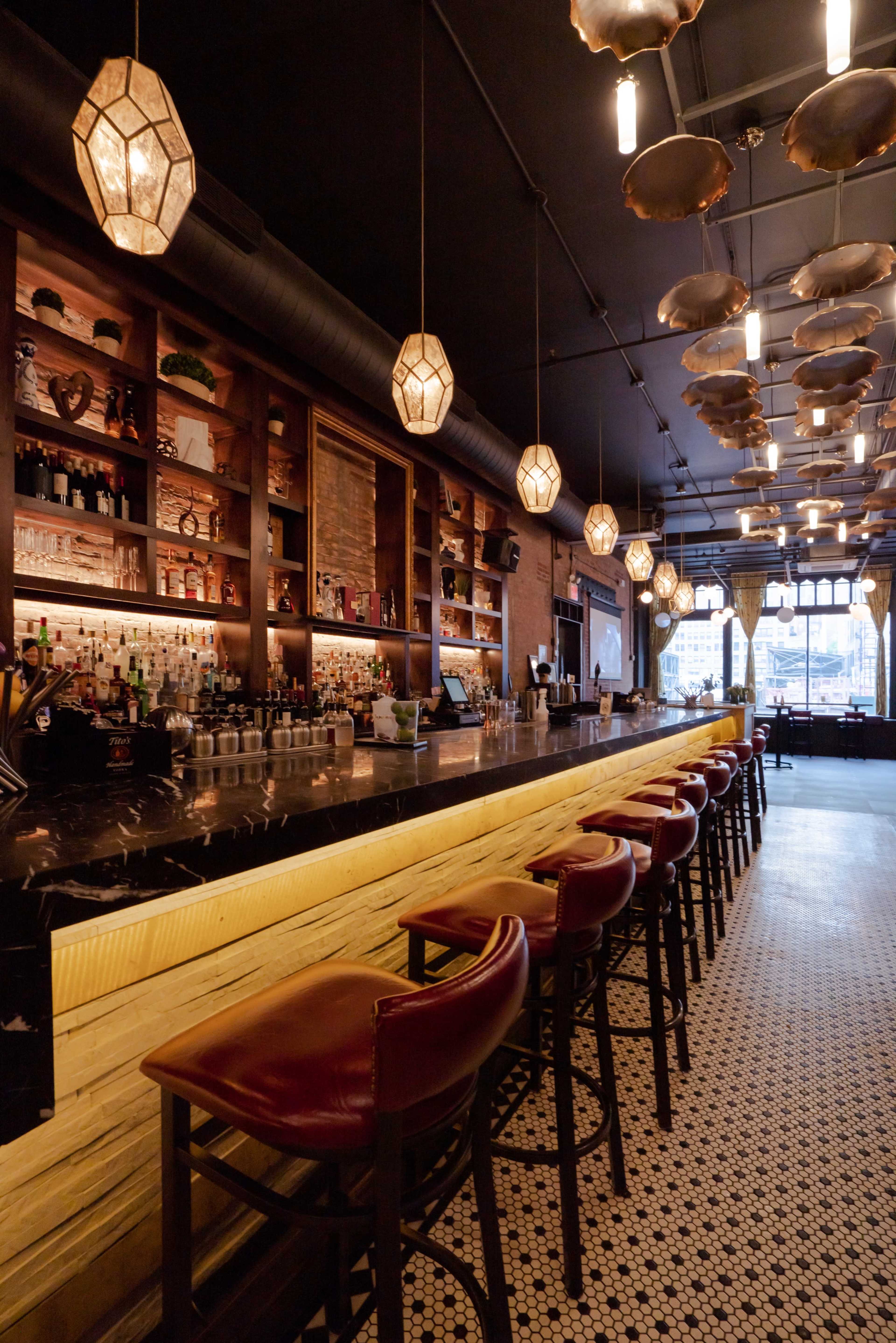 A modern bar interior with a long, shiny countertop, decorative light fixtures, and a well-stocked shelf behind the bar.