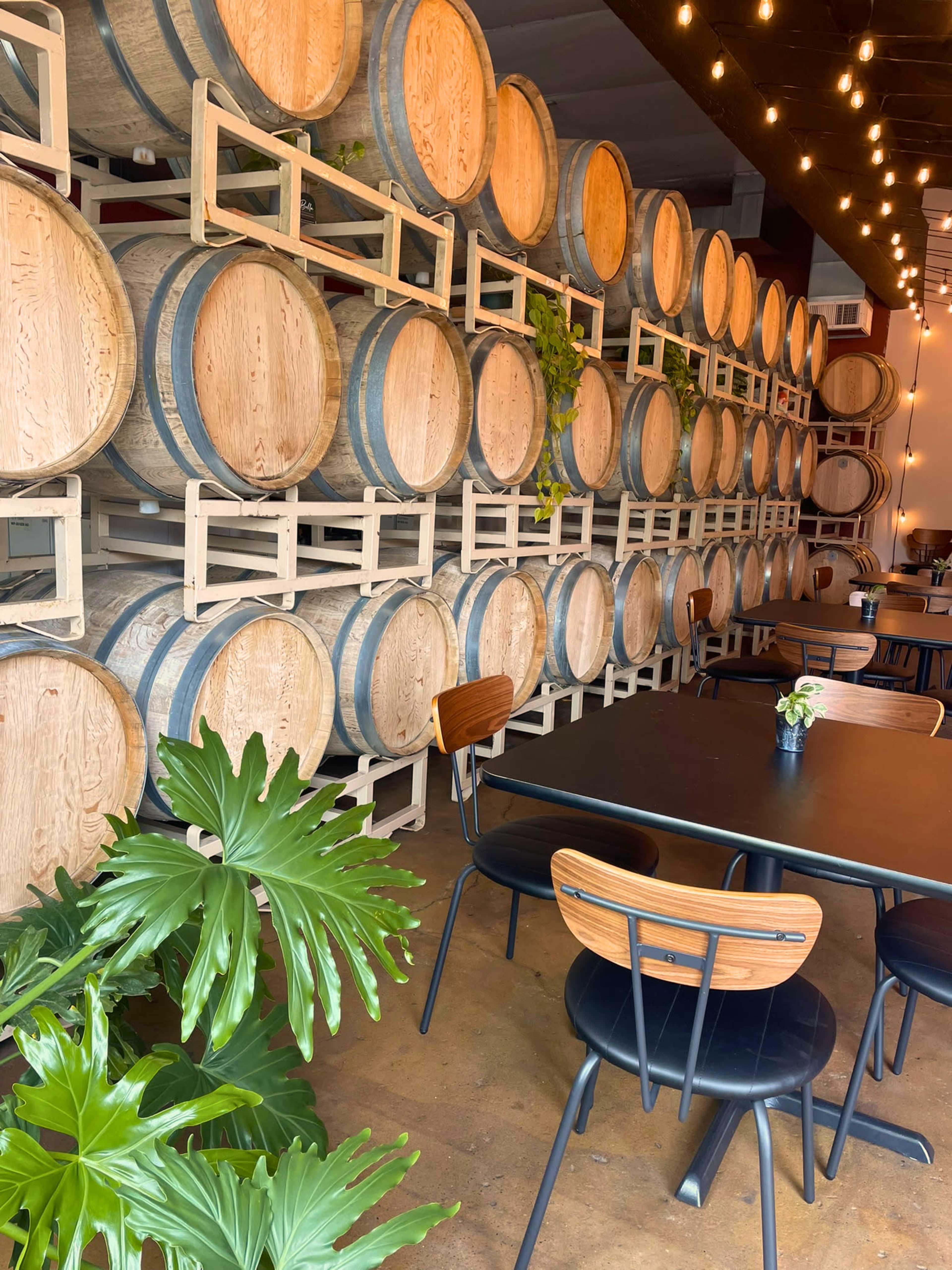 The image shows a restaurant interior featuring a wall of wooden barrels arranged on shelves, with tables and chairs positioned nearby under warm, hanging lights.
