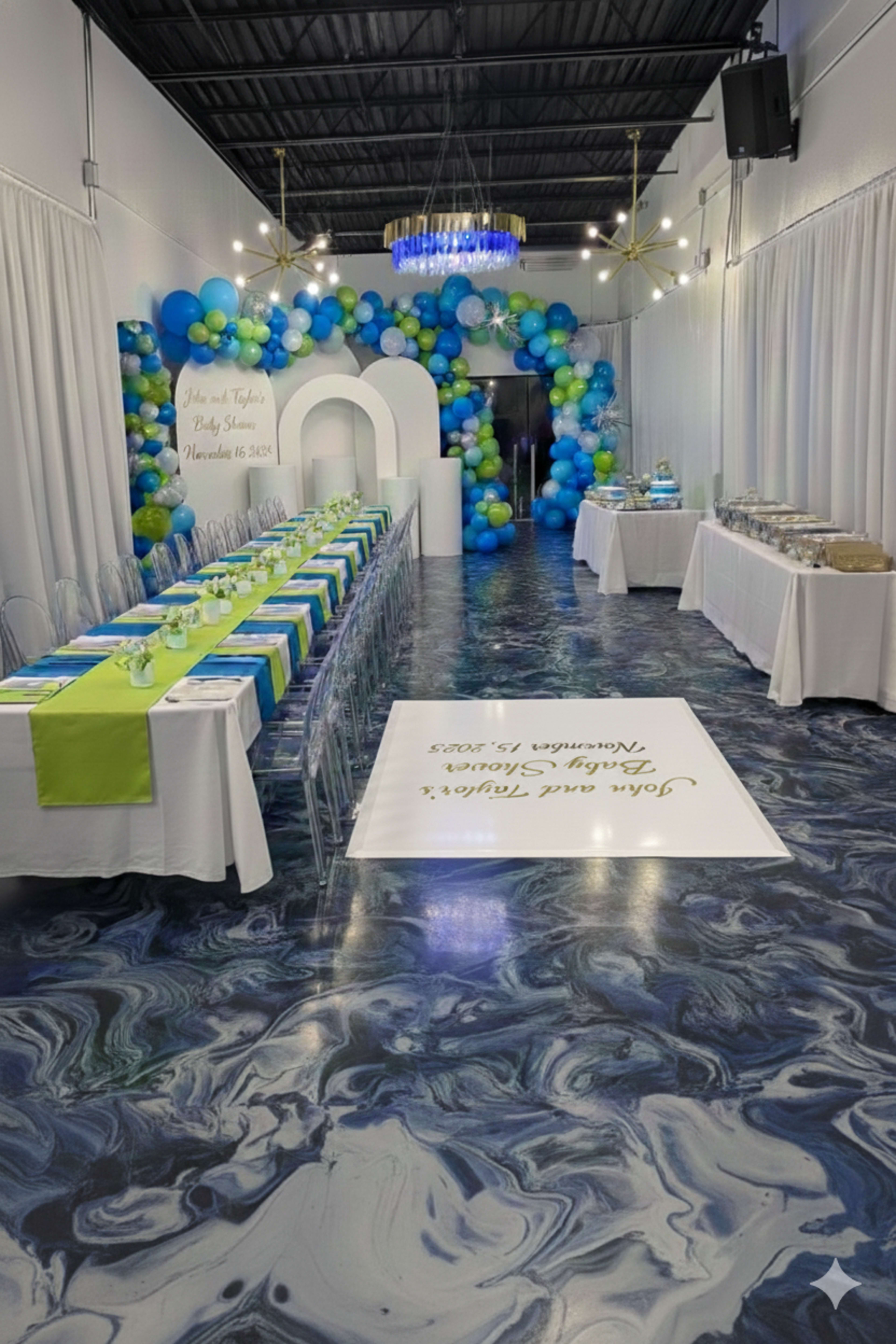 A decorated event space features a long table with blue and green table settings, colorful balloon garlands in the background, and a dessert display on the side.