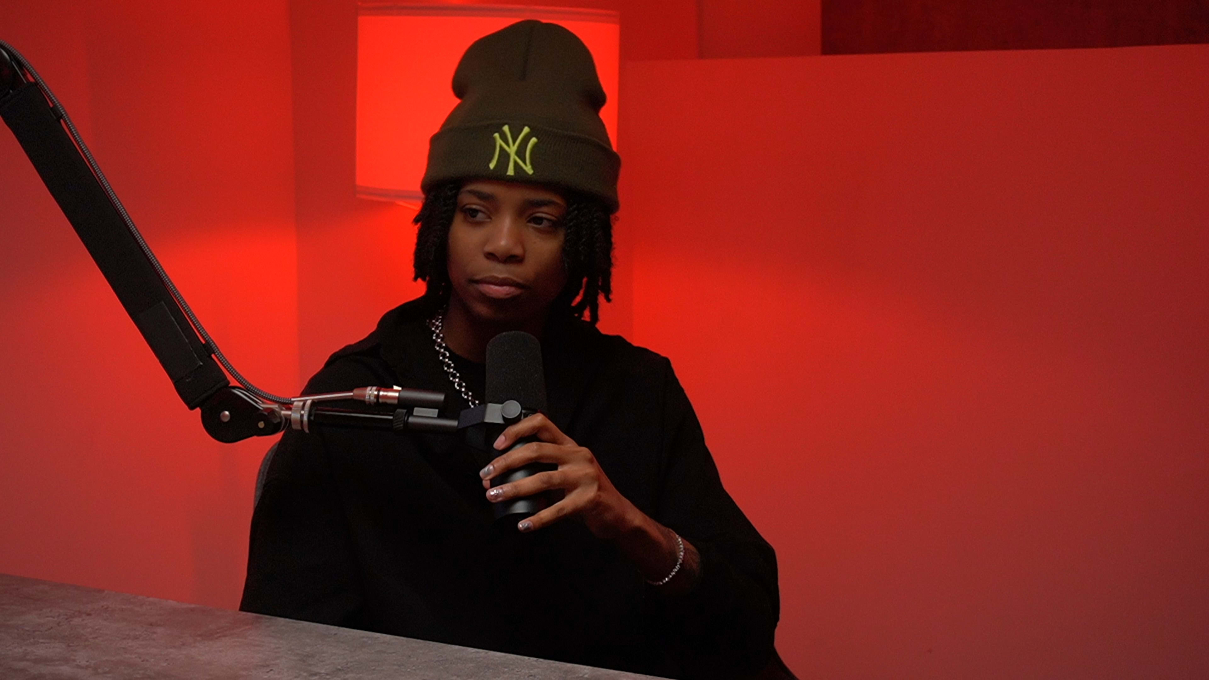 A person wearing a beanie and black clothing sits at a microphone in a studio with red lighting.