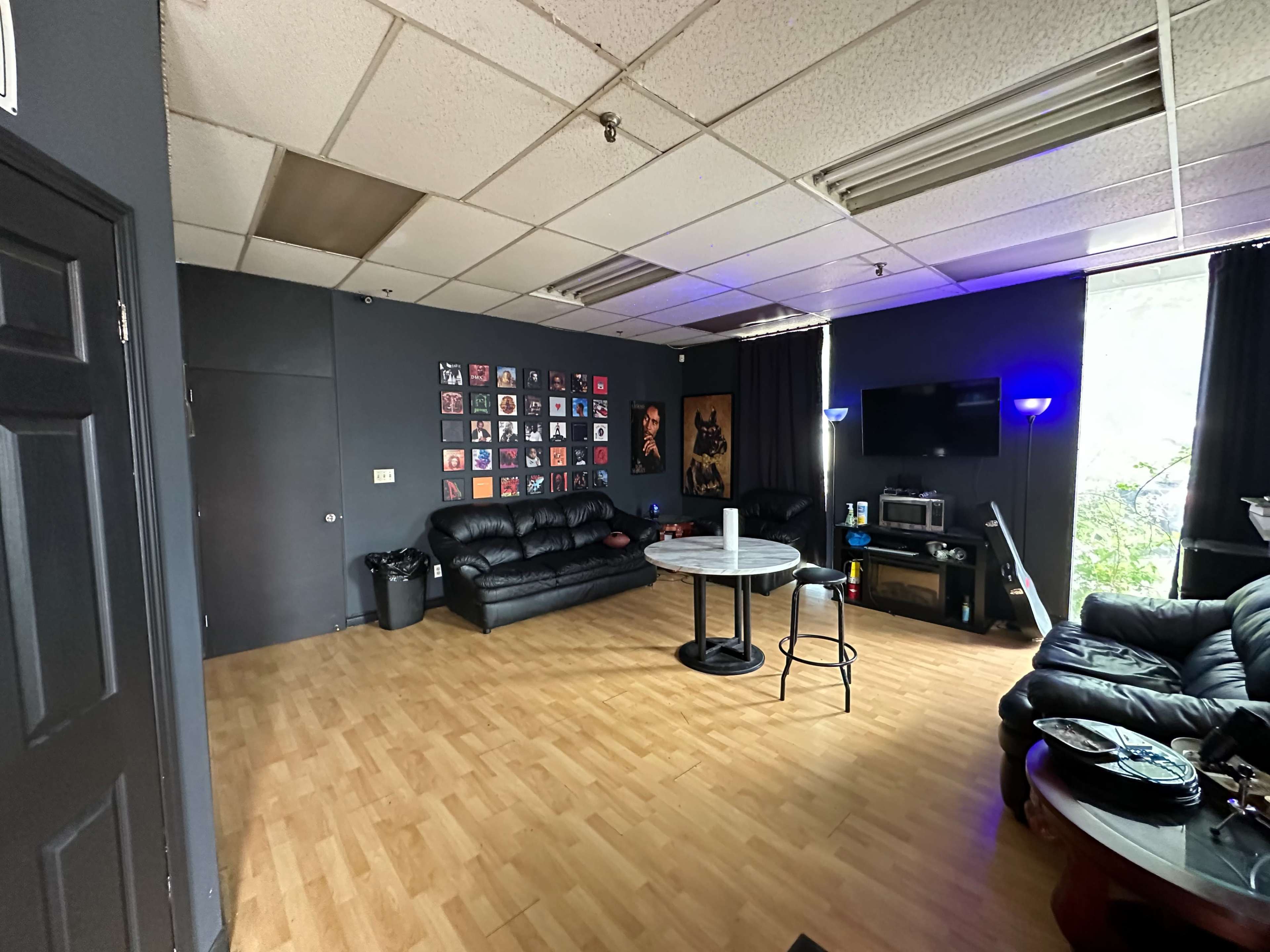 The image shows a room with a wooden floor, a black leather couch, a round table, a television, and a wall adorned with multiple framed pictures.