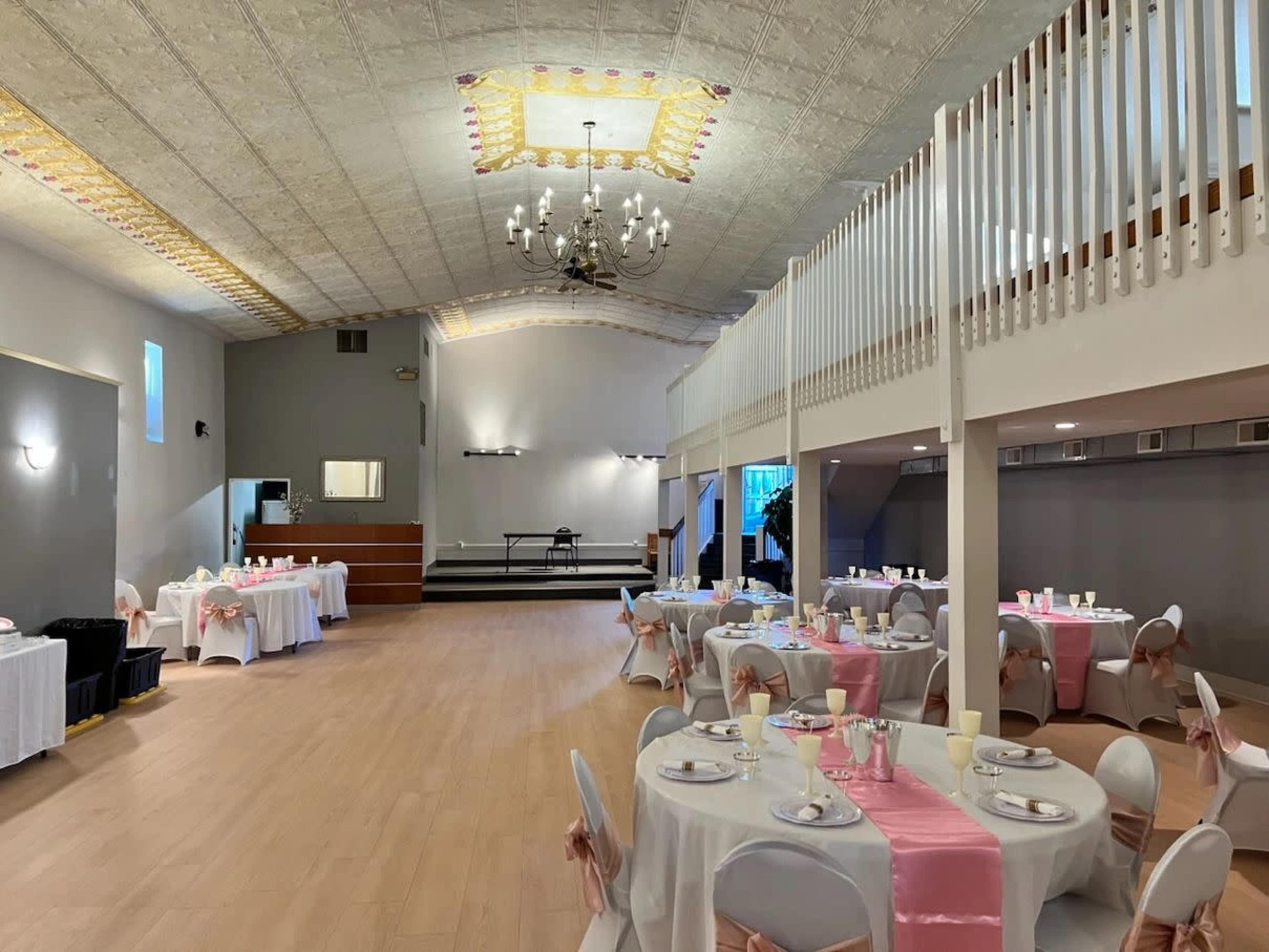 The image shows an elegant event space with round tables set for dining, each adorned with pink accents, and a raised stage area at the back.